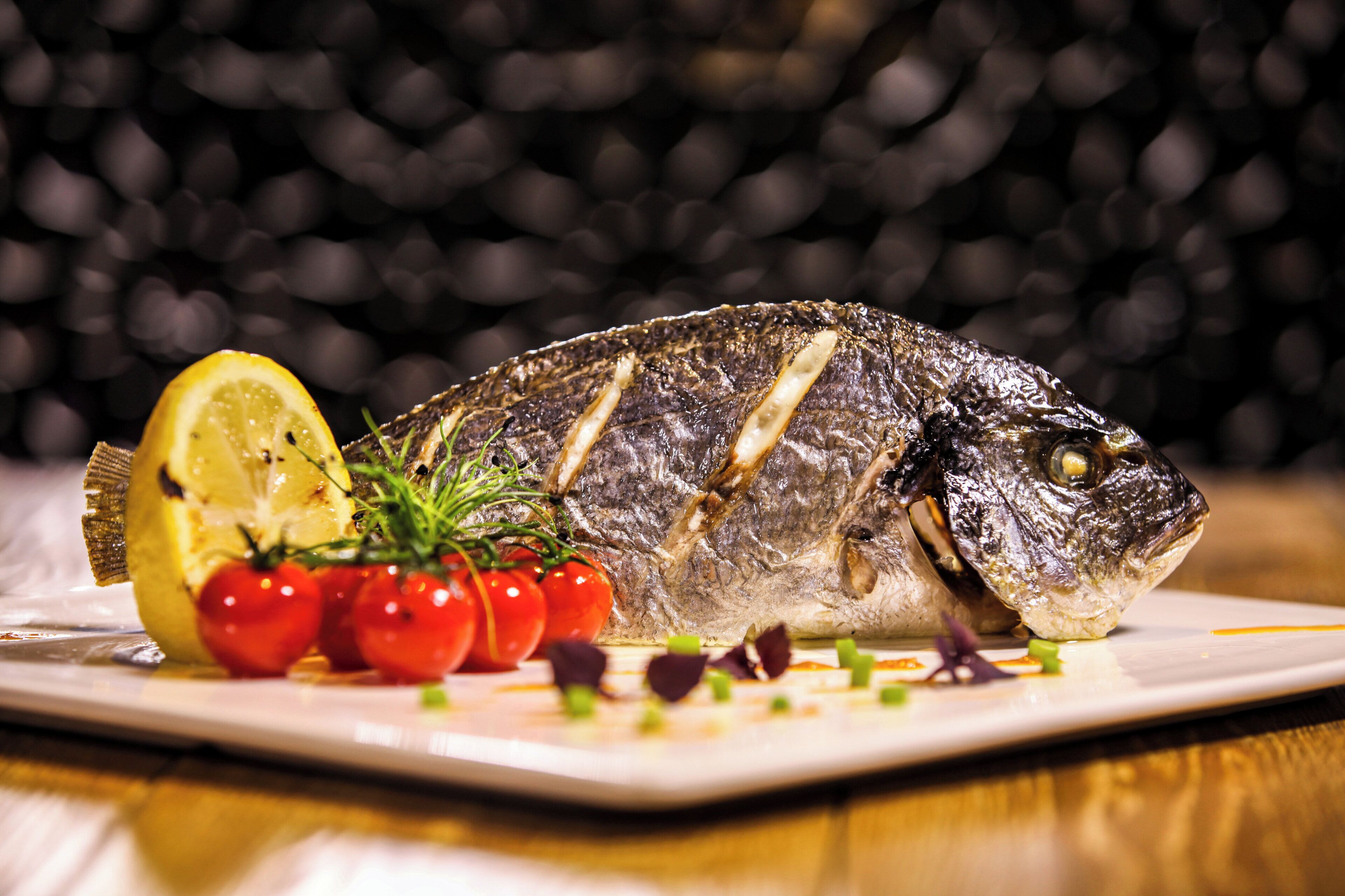 Un pescado entero a la parrilla en un plato con rodajas de limón, tomates cherry y hierbas.