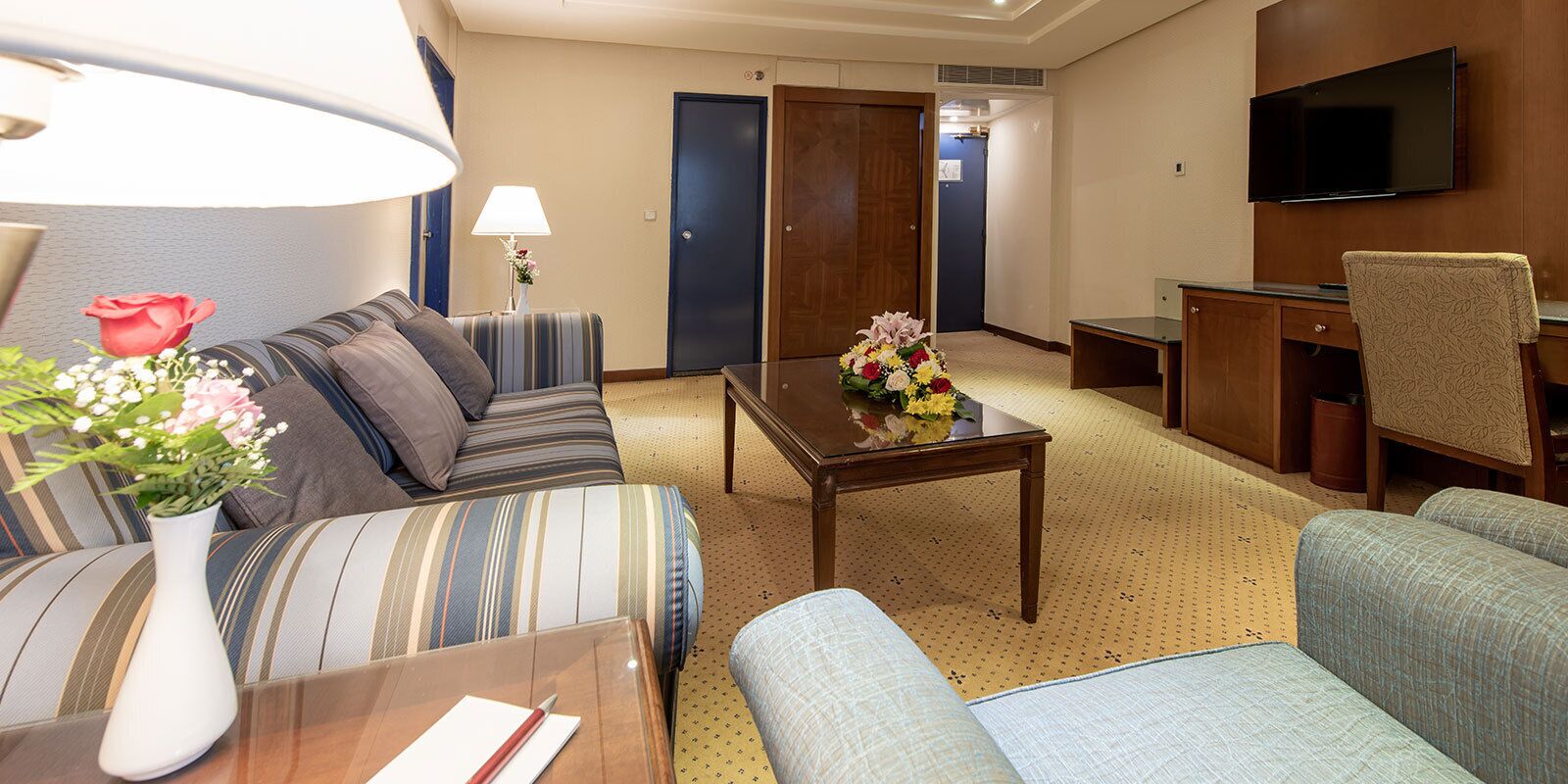 Une chambre d’hôtel comprenant un canapé, un fauteuil, une petite table avec un vase de fleurs, une lampe, un bureau avec une chaise, une télévision et une porte.