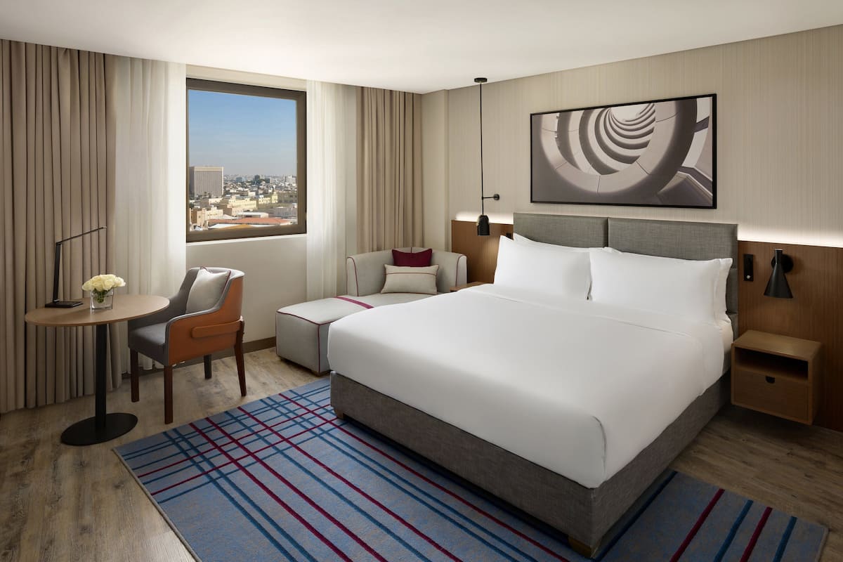 Une chambre d’hôtel avec un grand lit, un bureau avec une chaise, une petite table avec un vase, une vue sur la ville et une peinture abstraite moderne accrochée au mur.