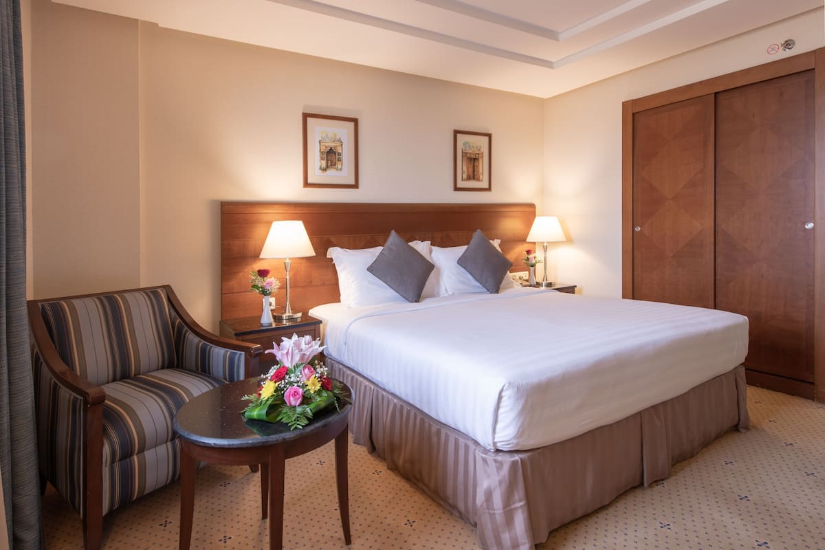 Une chambre d’hôtel avec un grand lit, deux tables de chevet avec des lampes, un canapé et une petite table décorée de fleurs.