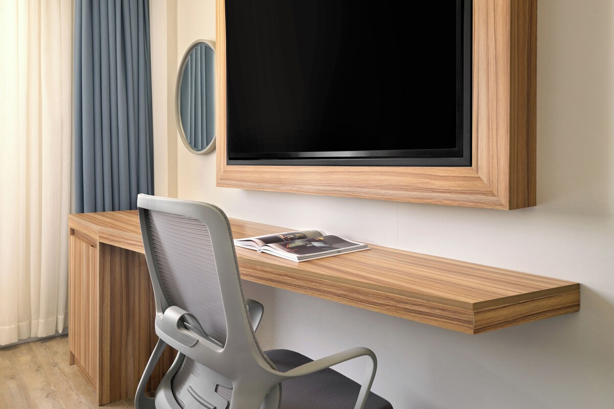 A modern office space with a wooden desk, a grey office chair, and a large flat-screen TV mounted on the wall.