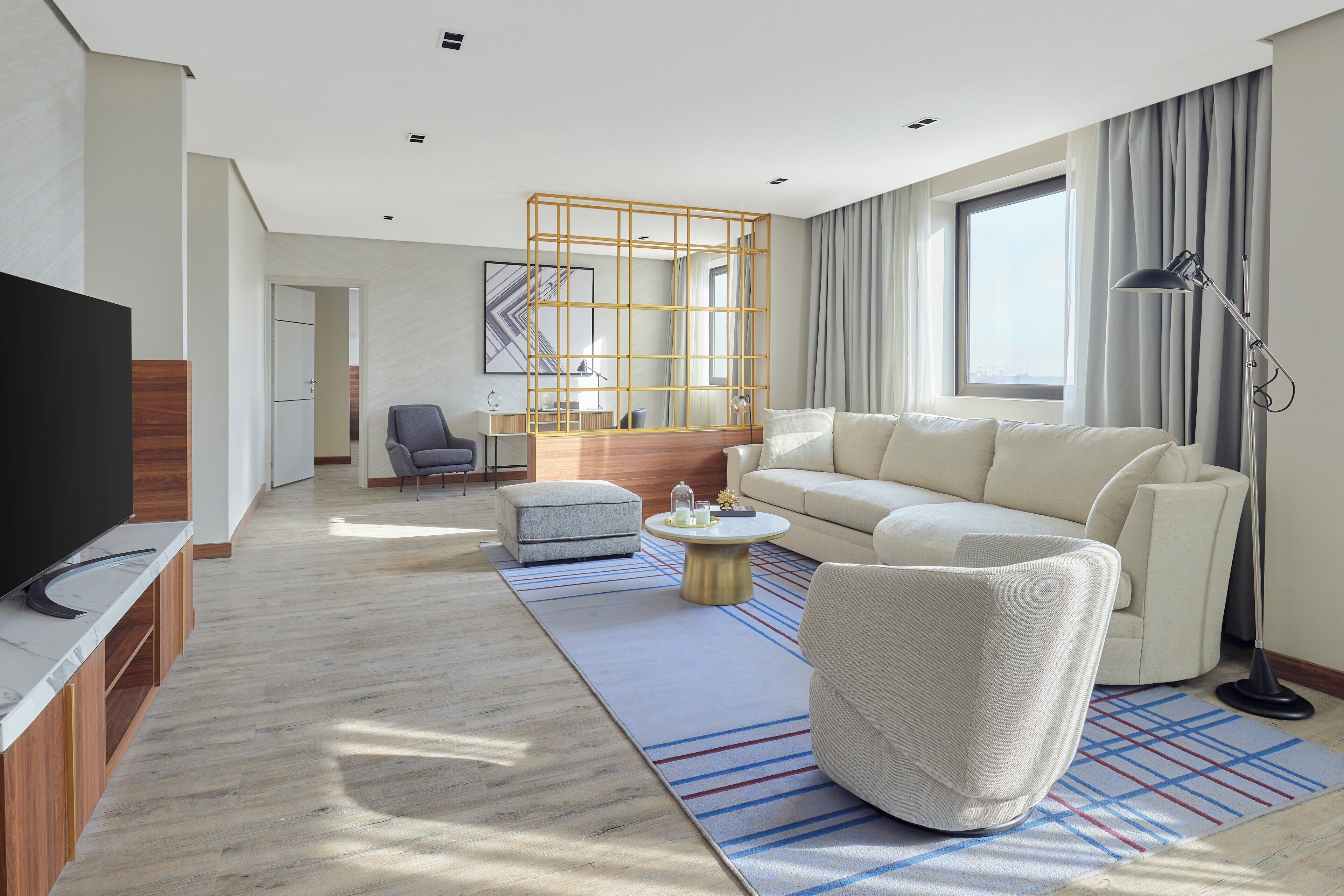A modern living room with a sofa, ottoman, and armchair, a flat-screen TV, a coffee table, and a large window with curtains.