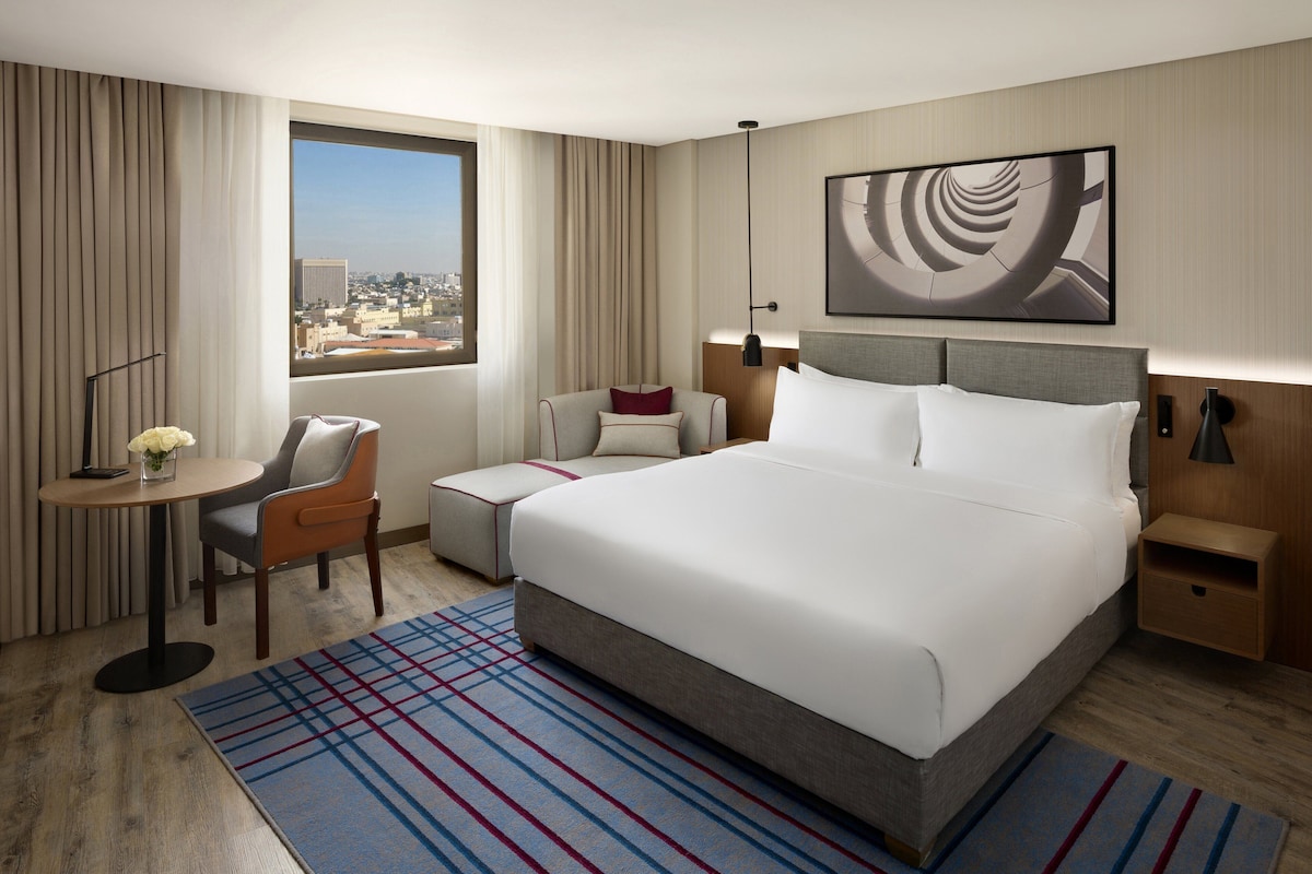 A hotel room with a large bed, a desk with a chair, a small table with a vase, a view of the city, and a modern abstract painting on the wall.