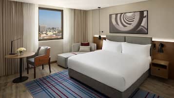 A hotel room with a large bed, a desk with a chair, a small table with a vase, a view of the city, and a modern abstract painting on the wall.