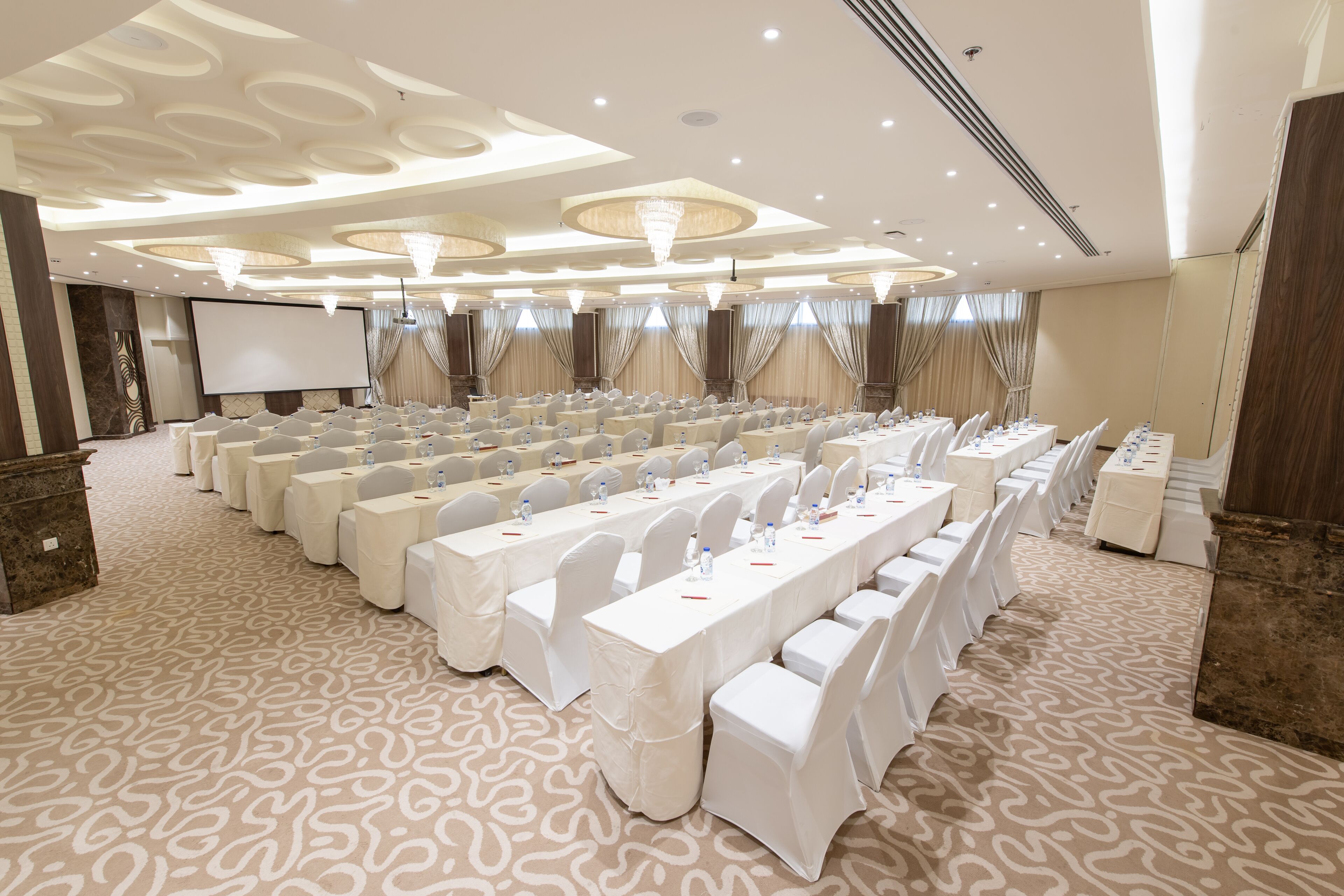 Un salón de conferencias con filas de mesas, sillas y una pantalla de proyección.