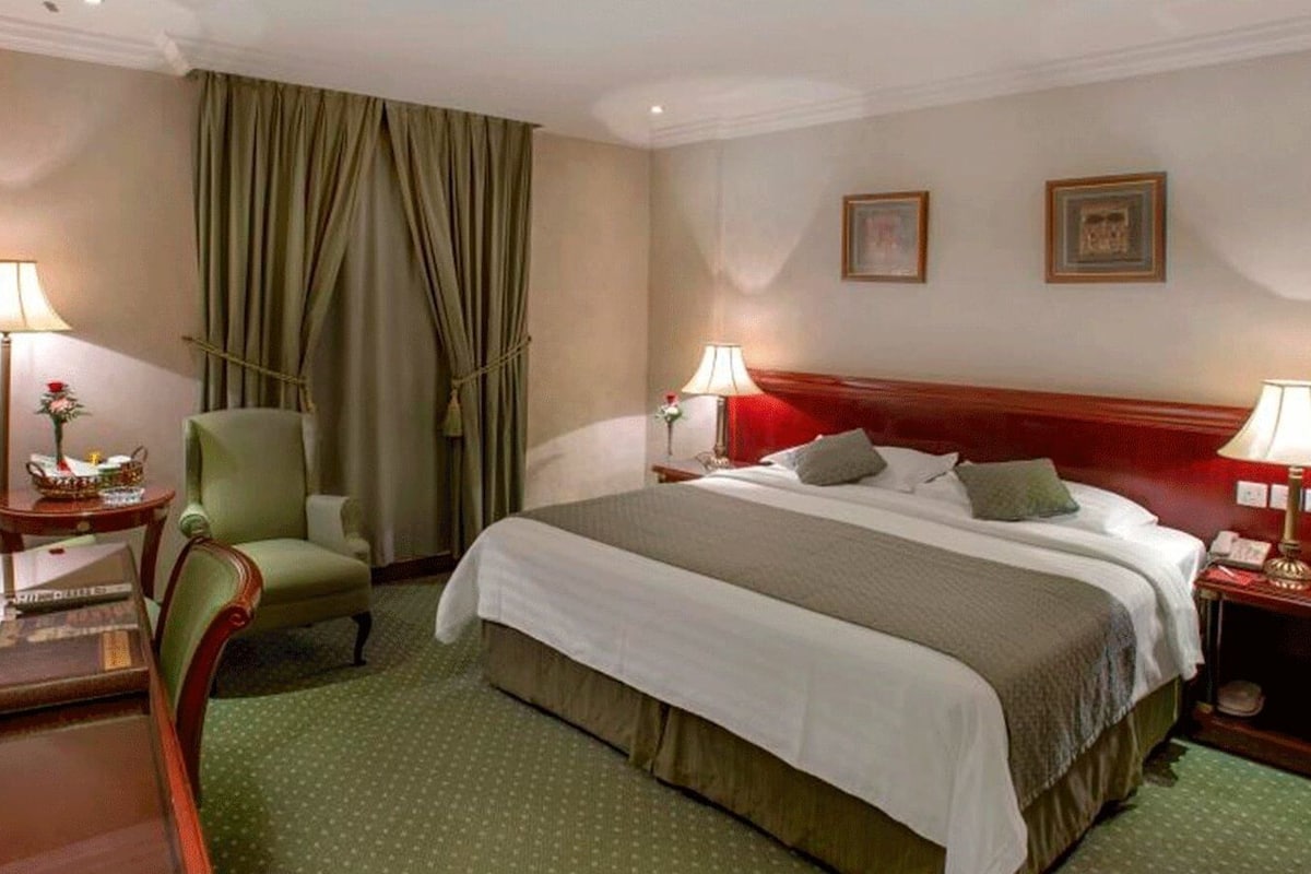 A hotel room with a bed, two bedside tables, a chair, a desk, a lamp, a mirror, and a window with curtains.