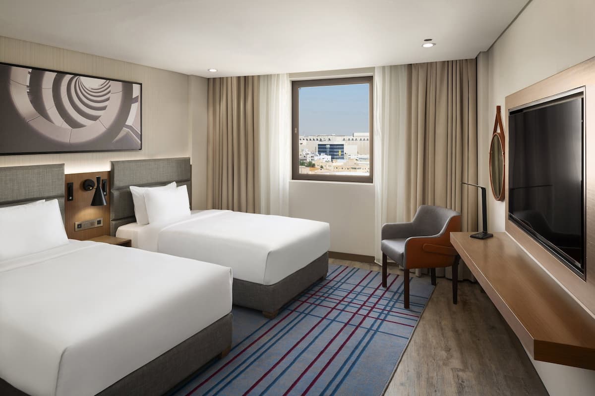 Une chambre d’hôtel avec deux lits, un bureau, une télévision et une grande fenêtre donnant sur la ville.