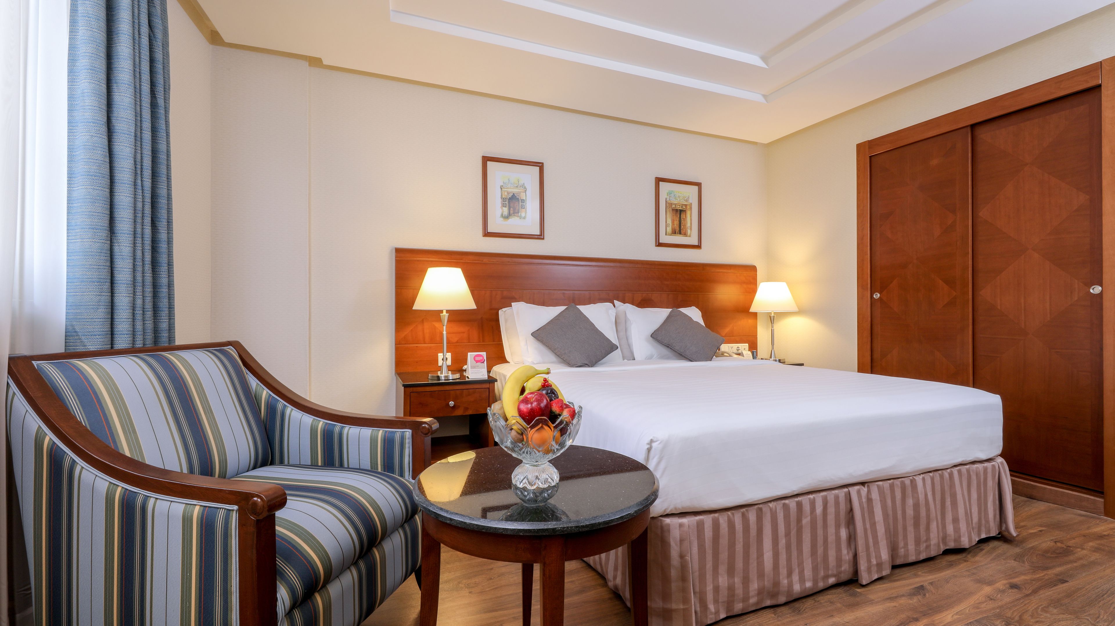 A hotel room with a bed, a chair, a small table with a fruit bowl, two wall-mounted lamps, and a wooden wardrobe.
