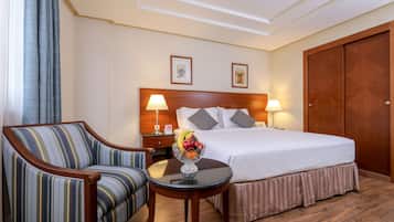 A hotel room with a bed, a chair, a small table with a fruit bowl, two wall-mounted lamps, and a wooden wardrobe.