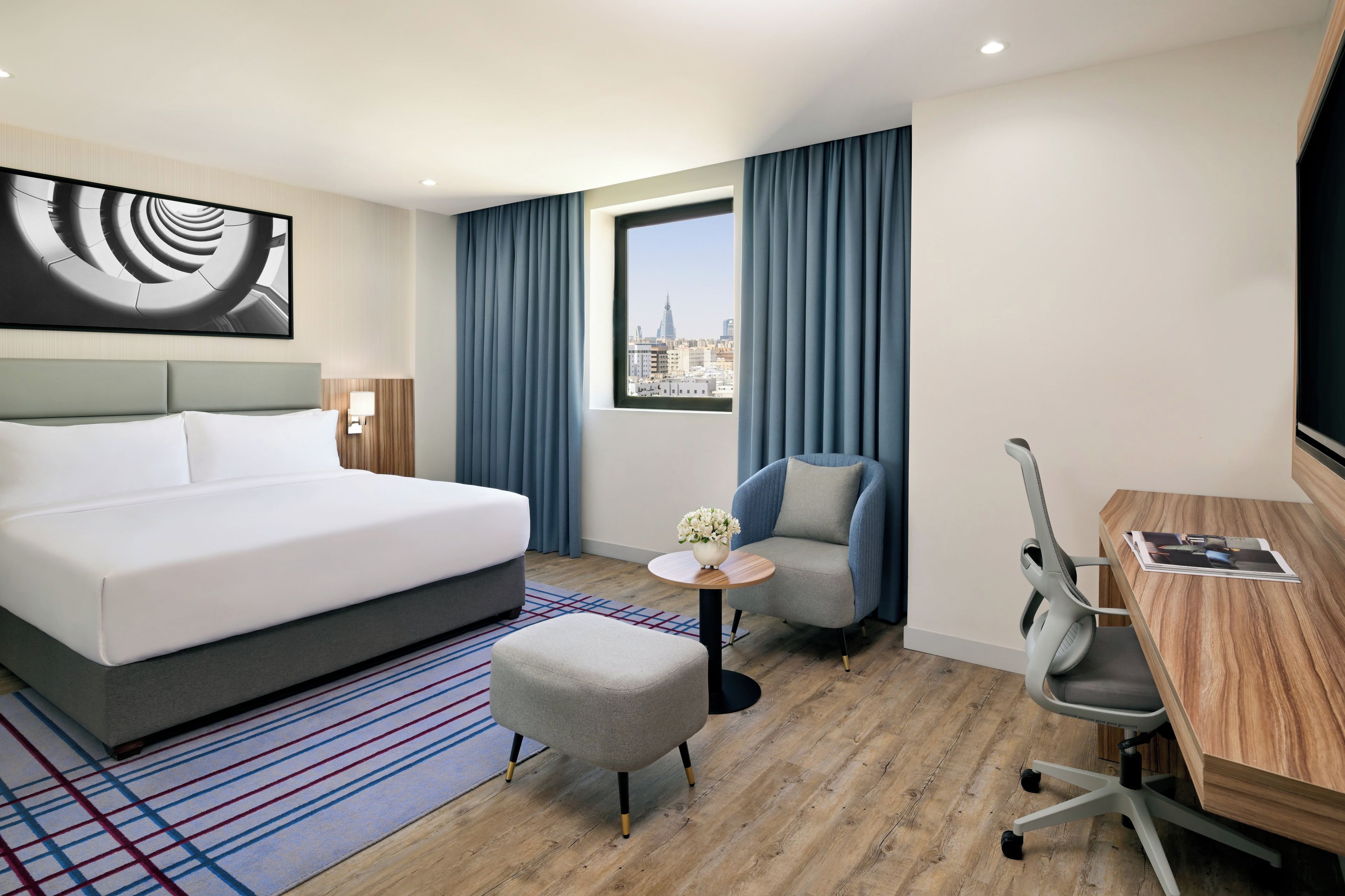 A hotel room with a large bed, a desk with a chair, a small table with a vase, and a view of a cityscape through a window.