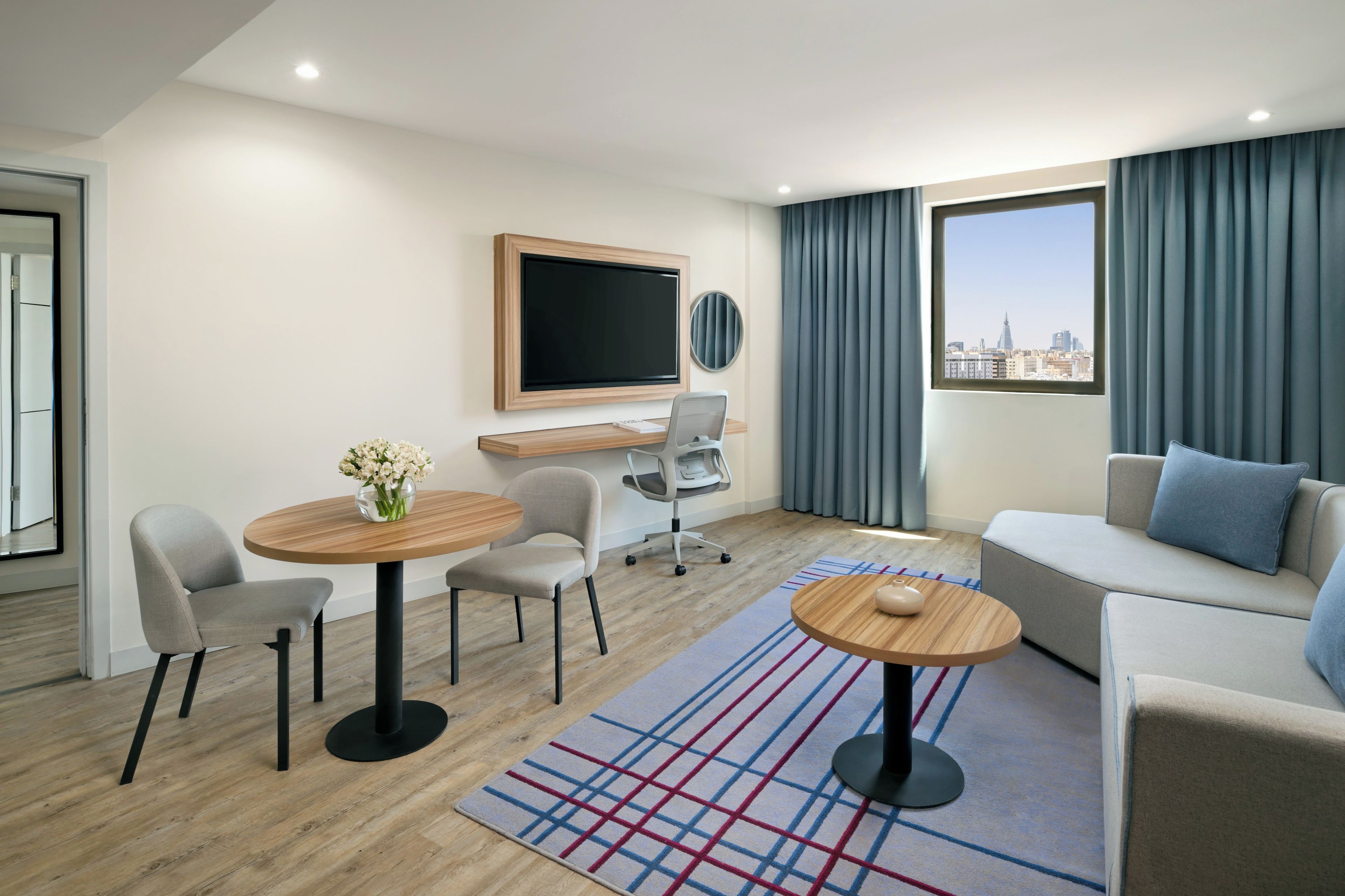 A modern hotel room with a flat-screen TV, a sofa, a dining table with chairs, a small desk with a chair, and a view of the city through large windows.
