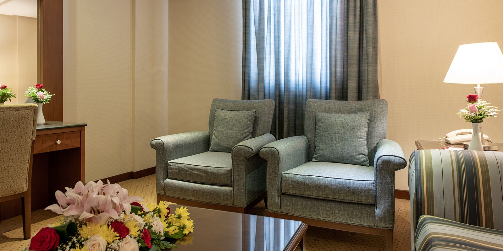 Une chambre d’hôtel avec deux fauteuils gris, une petite table sur laquelle se trouve un vase de fleurs, une lampe et un téléphone posé sur une table d’appoint.