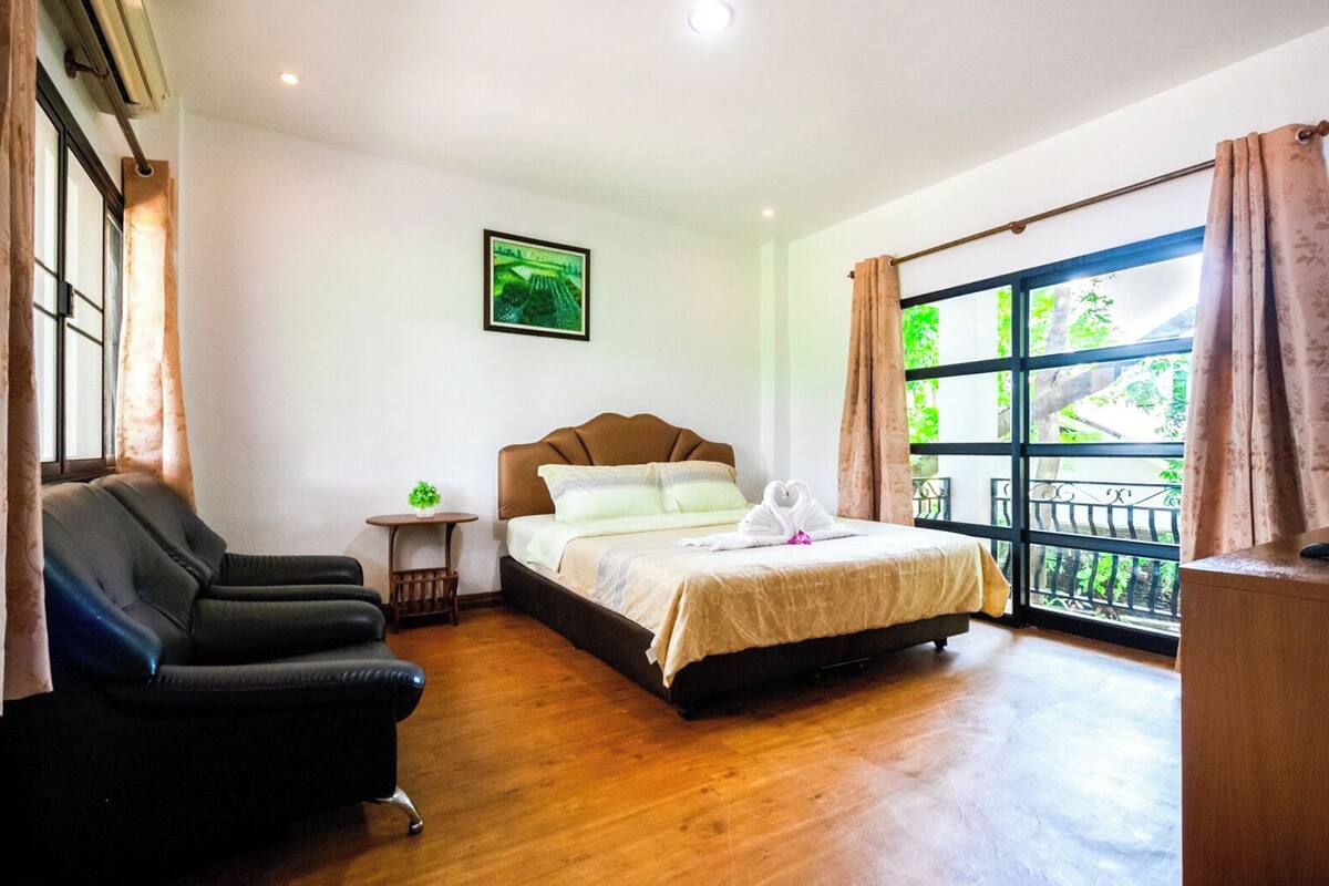 A hotel room with a bed, a desk, a chair, a small table with a plant, and a large window with a view of greenery.