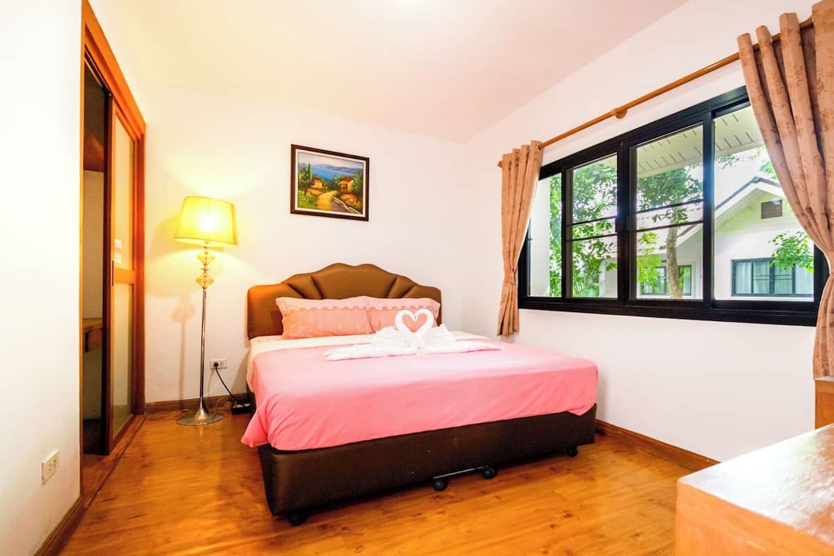 A neatly arranged bedroom with a bed, a bedside lamp, a window with curtains, and a painting on the wall.