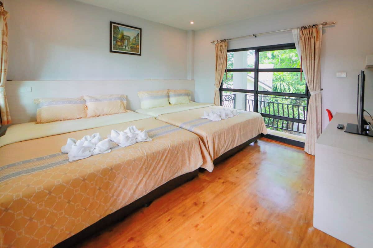 A hotel room with two beds, a balcony with a view of greenery, a painting on the wall, and a desk with a computer.