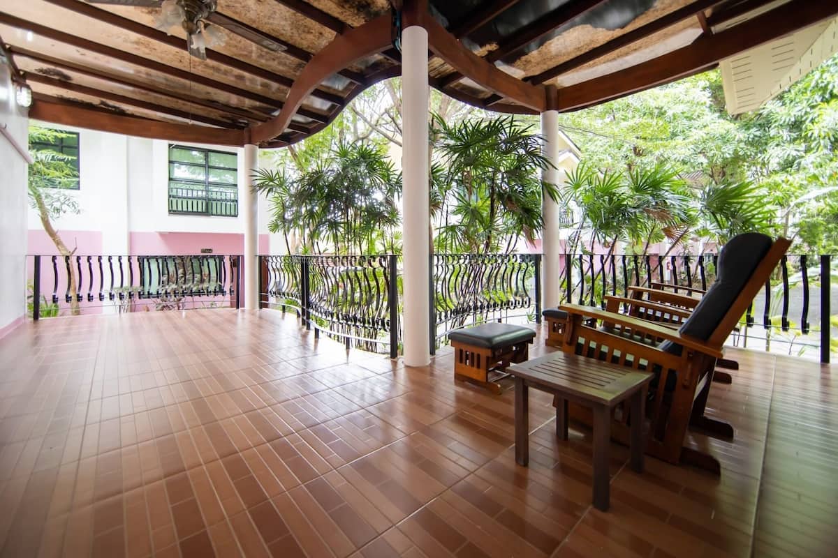 A covered outdoor area with wooden flooring, a table, and chairs, surrounded by a metal railing and greenery.