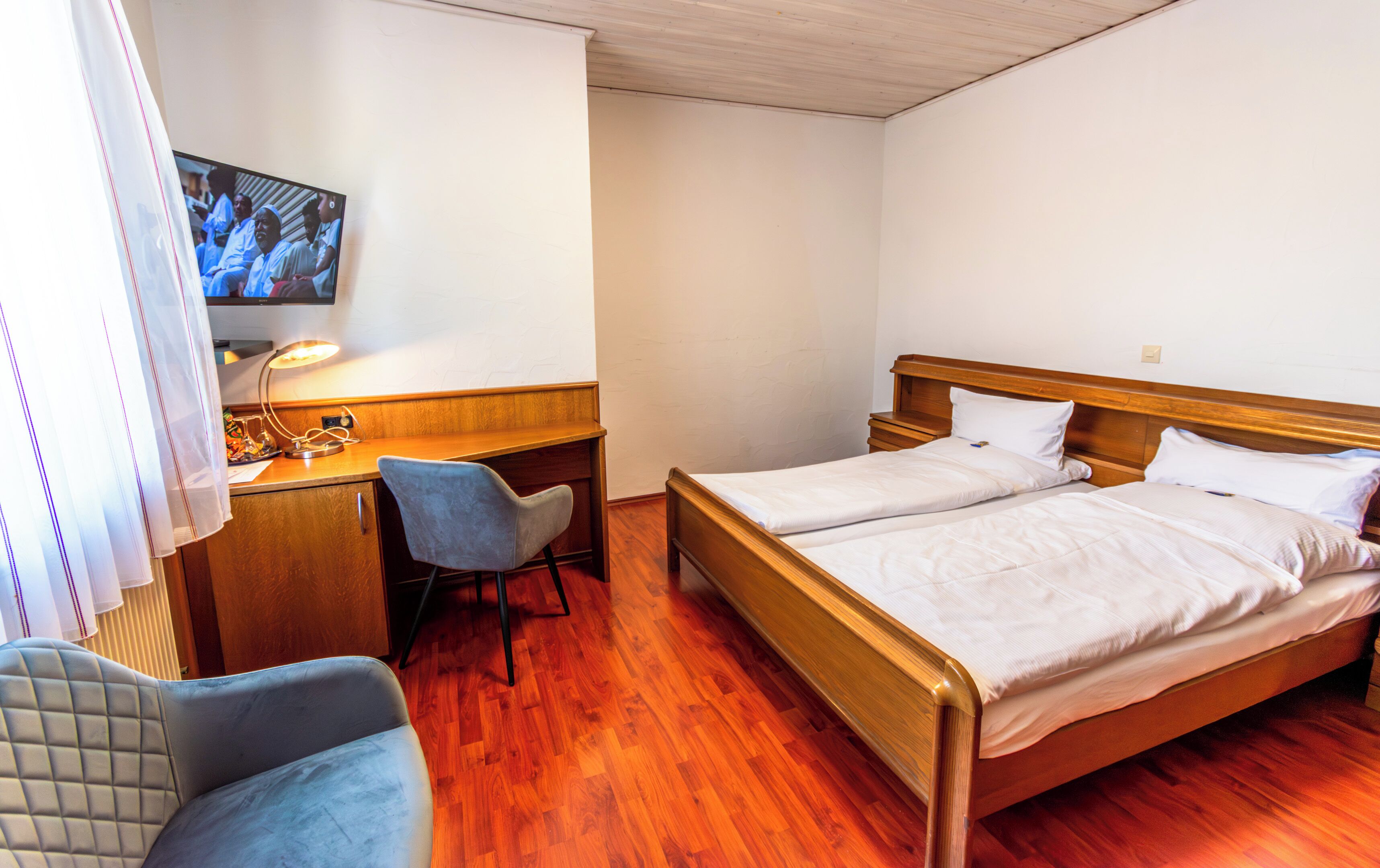 Ein Hotelzimmer mit einem Holzbett, einem Schreibtisch mit Stuhl, einem Fernseher und einem Fenster mit Vorhängen.