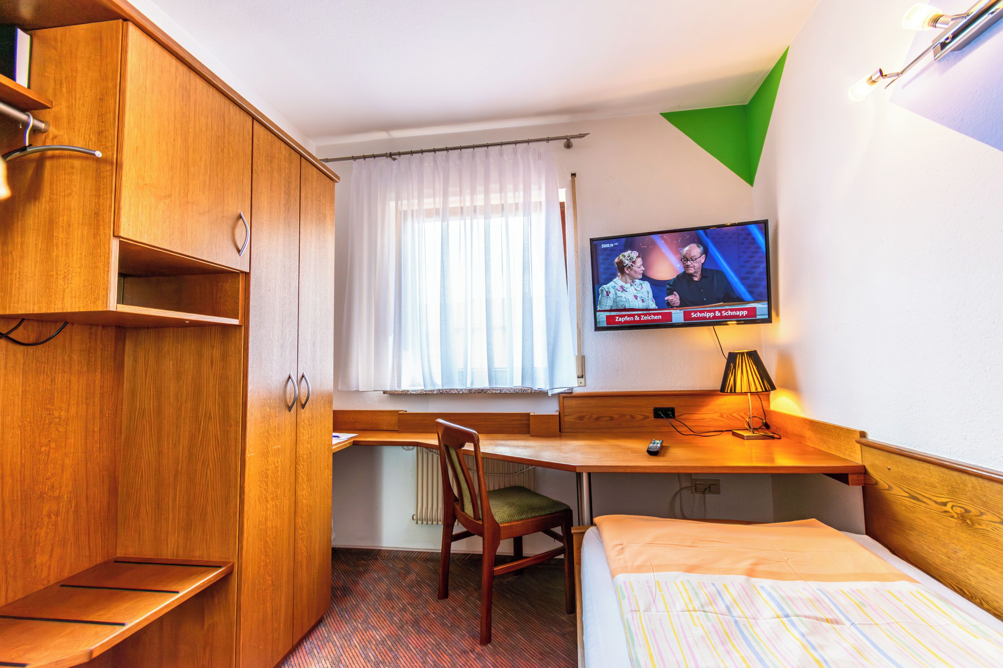 A hotel room with a wooden desk, a television mounted on the wall, a bed with a colorful striped bedspread, and a window with white curtains.