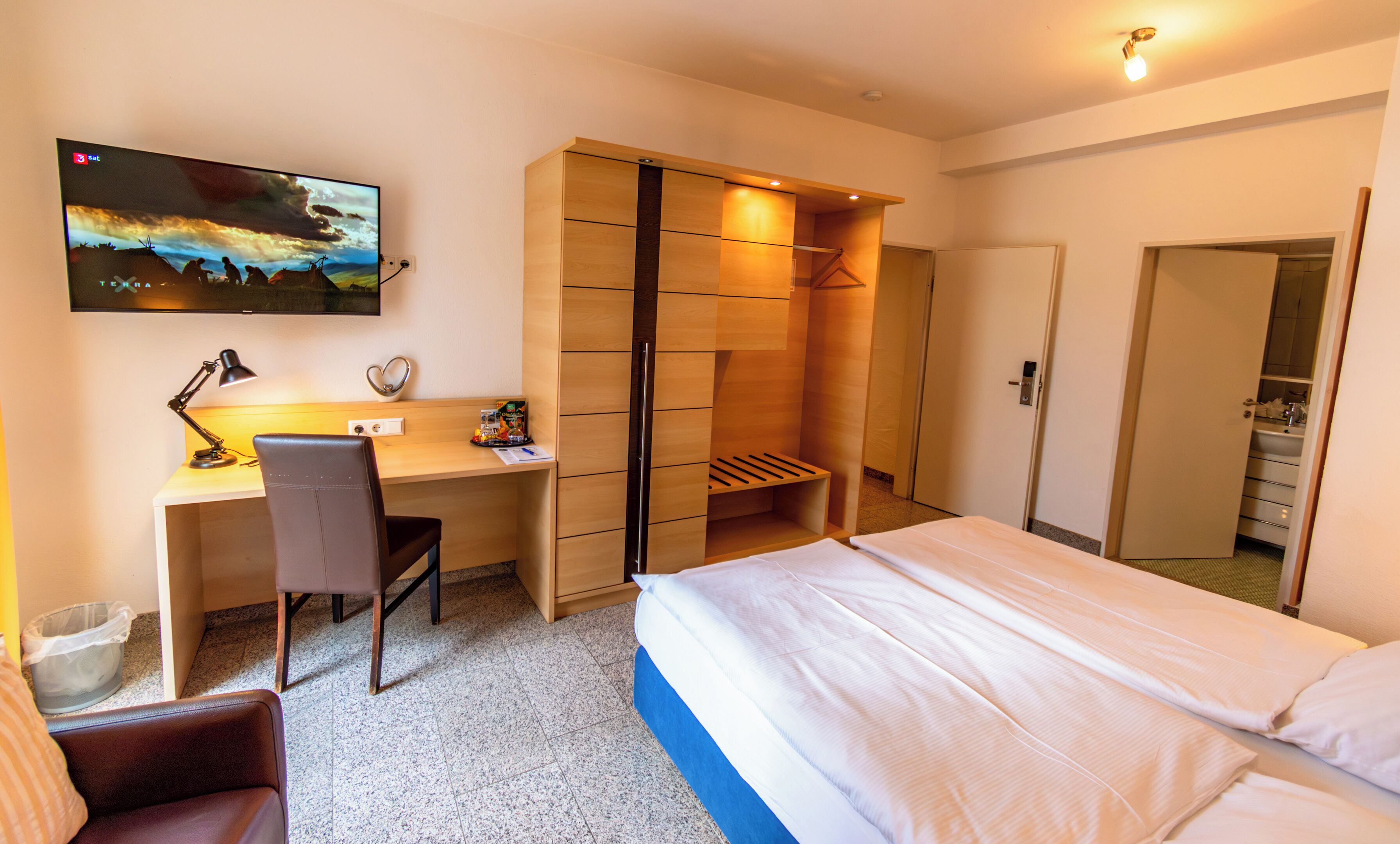 A hotel room with a bed, a desk with a chair, a TV mounted on the wall, and a wardrobe.