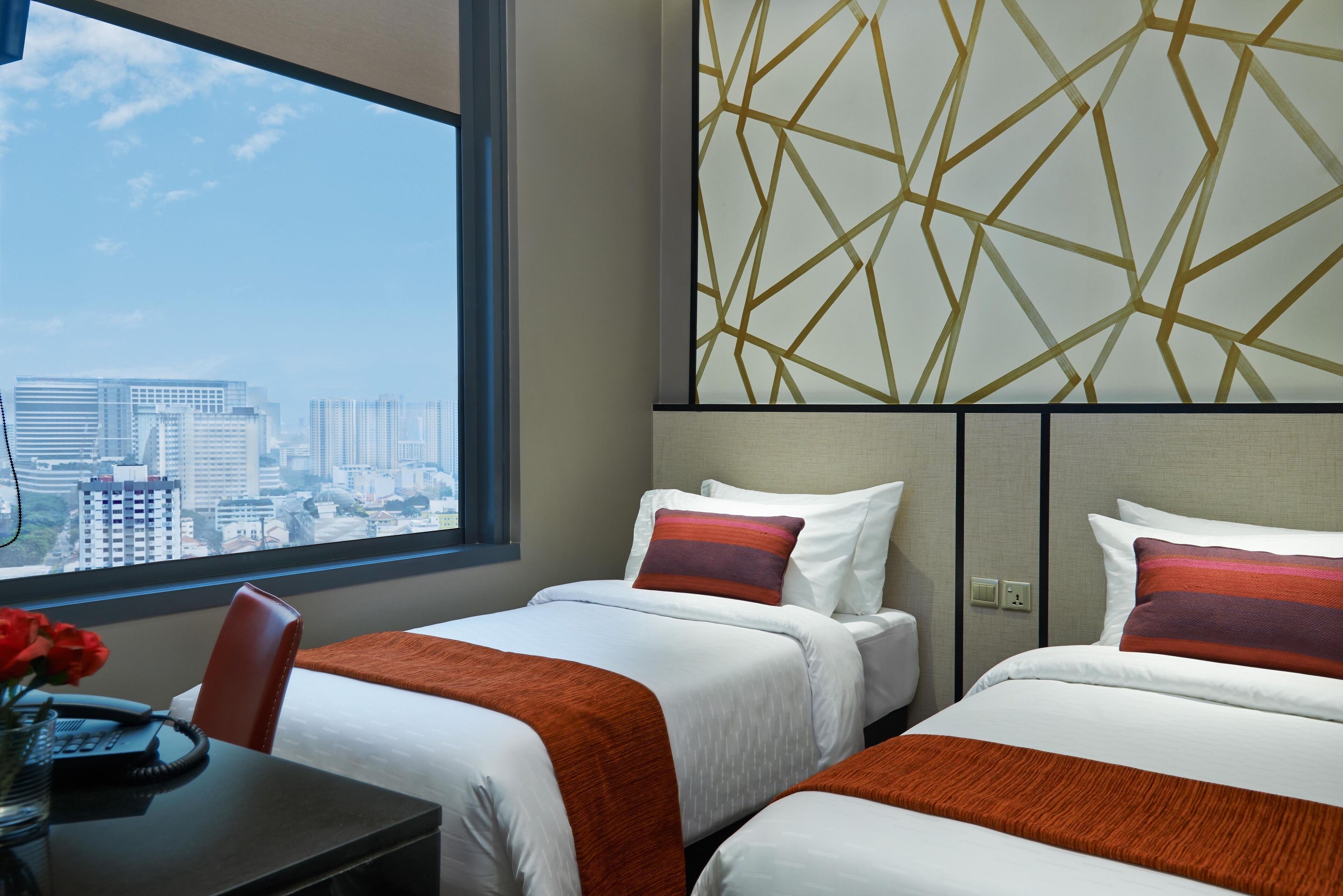 A hotel room with two beds, a desk with a telephone, and a view of a cityscape through a large window.