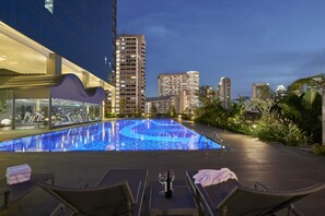 A rooftop pool with lounge chairs and a table, surrounded by modern buildings and greenery.