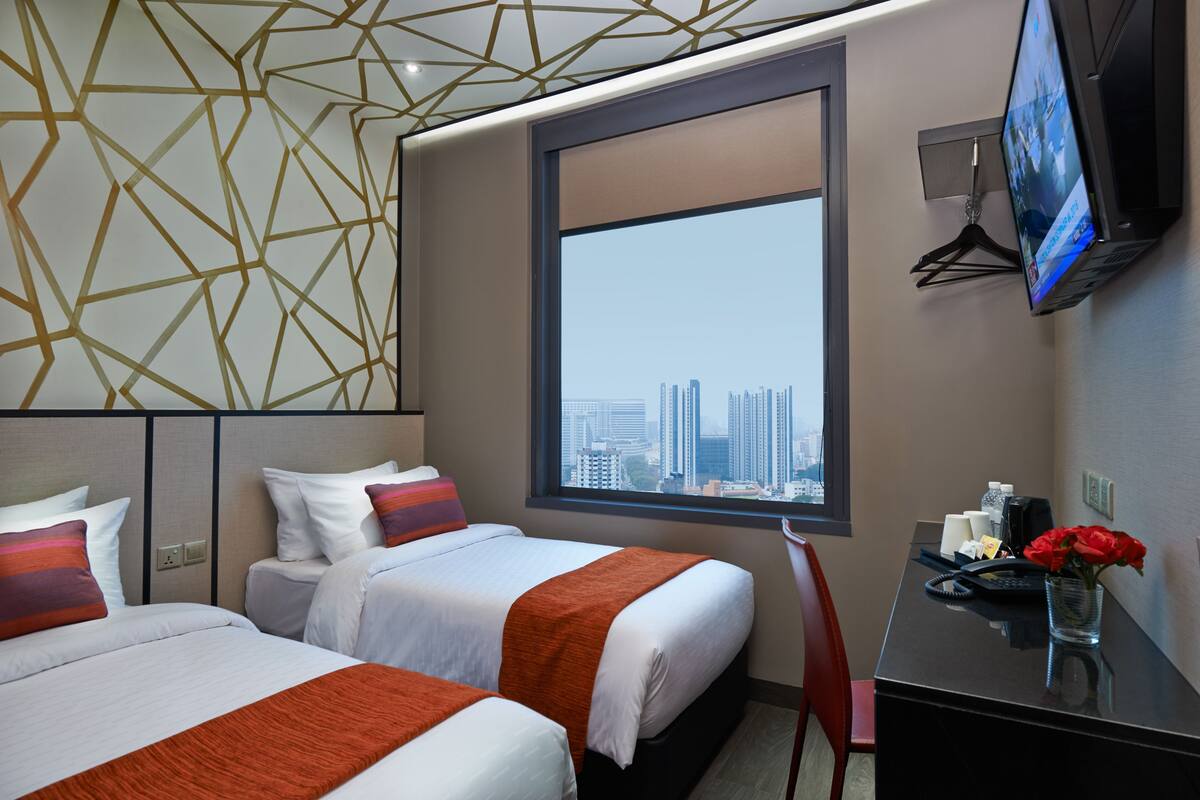A hotel room with two beds, a large window with a city view, a desk with a phone and a vase of flowers, and a TV mounted on the wall.