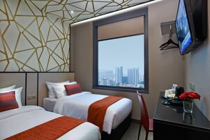 A hotel room with two beds, a large window with a city view, a desk with a phone and a vase of flowers, and a TV mounted on the wall.