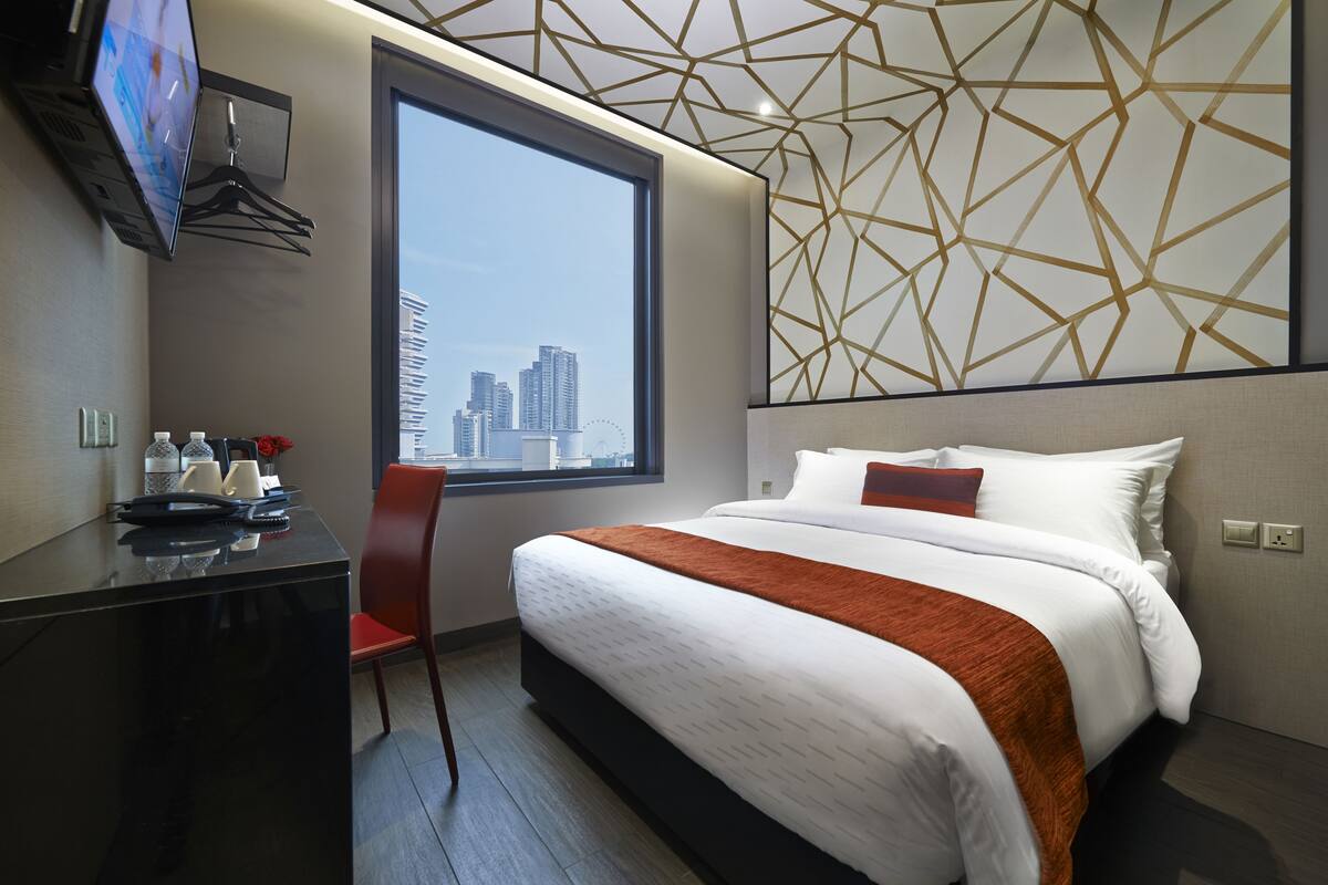 A hotel room with a large bed, a desk with a chair, a TV mounted on the wall, and a window with a city view.