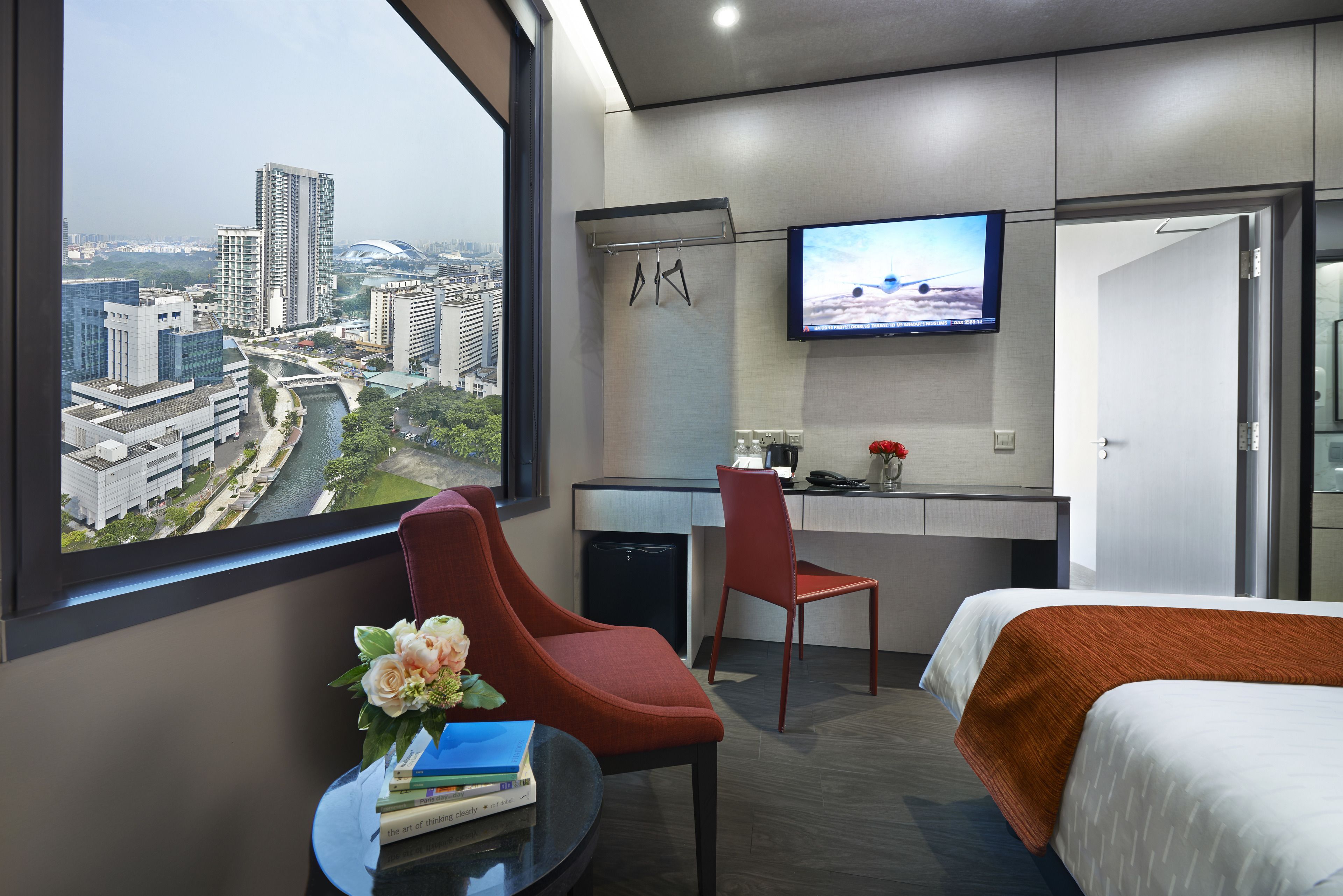 A hotel room with a large window offering a city view, a desk with a chair, a bed, and a TV mounted on the wall.