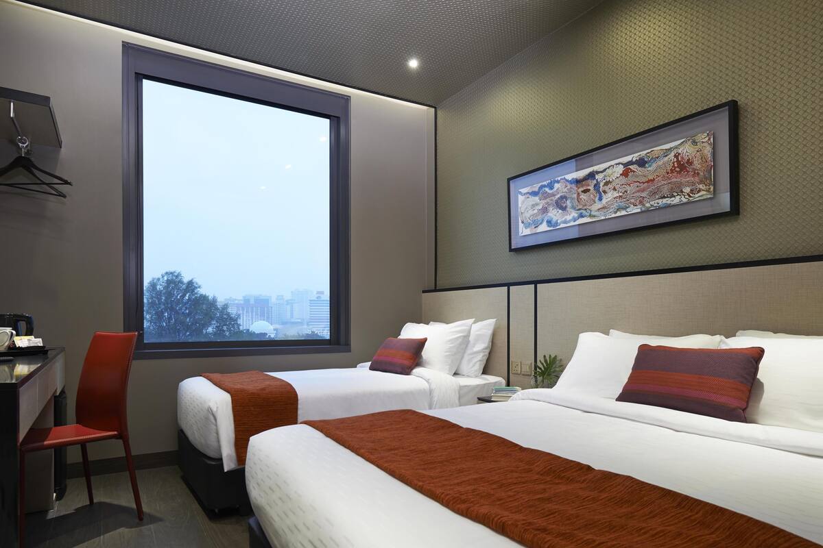 A hotel room with two beds, a desk, a chair, a large window, and a framed artwork on the wall.