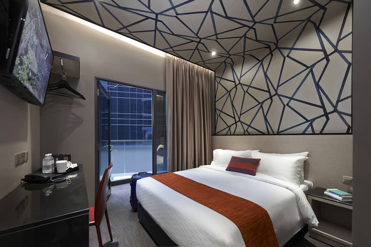 A hotel room with a bed, a desk with a telephone, a chair, a TV mounted on the wall, and a window with curtains.