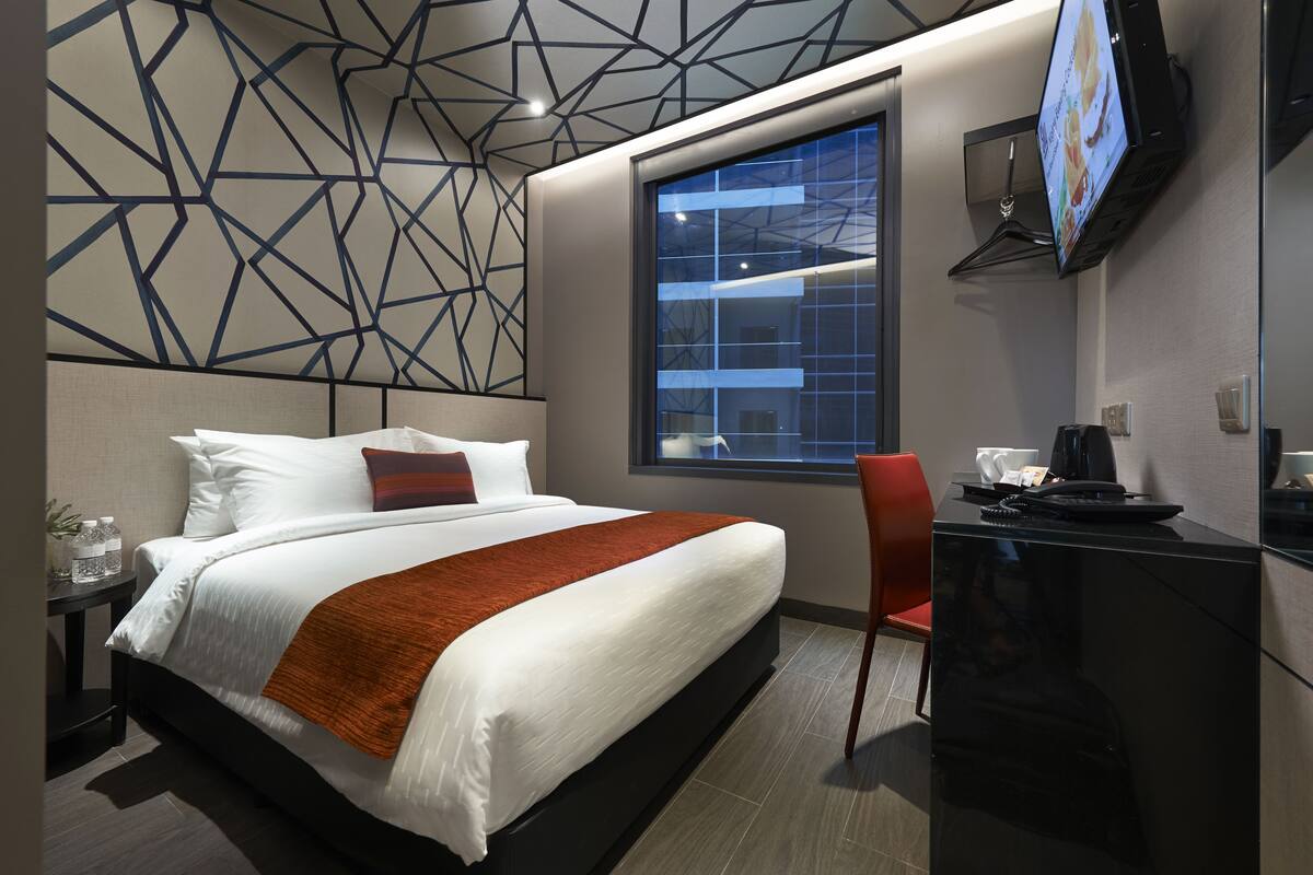 A modern hotel room with a large bed, a desk, a TV, and a window with a city view.