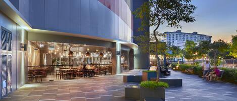 A modern building with a restaurant on the ground floor, outdoor seating, and a tree in the center.