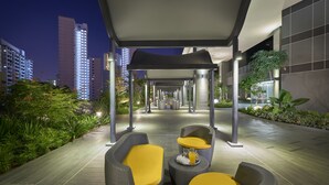 A covered outdoor seating area with wicker furniture and yellow cushions, surrounded by greenery and modern buildings in the background. - Hotel Boss (Singapore)