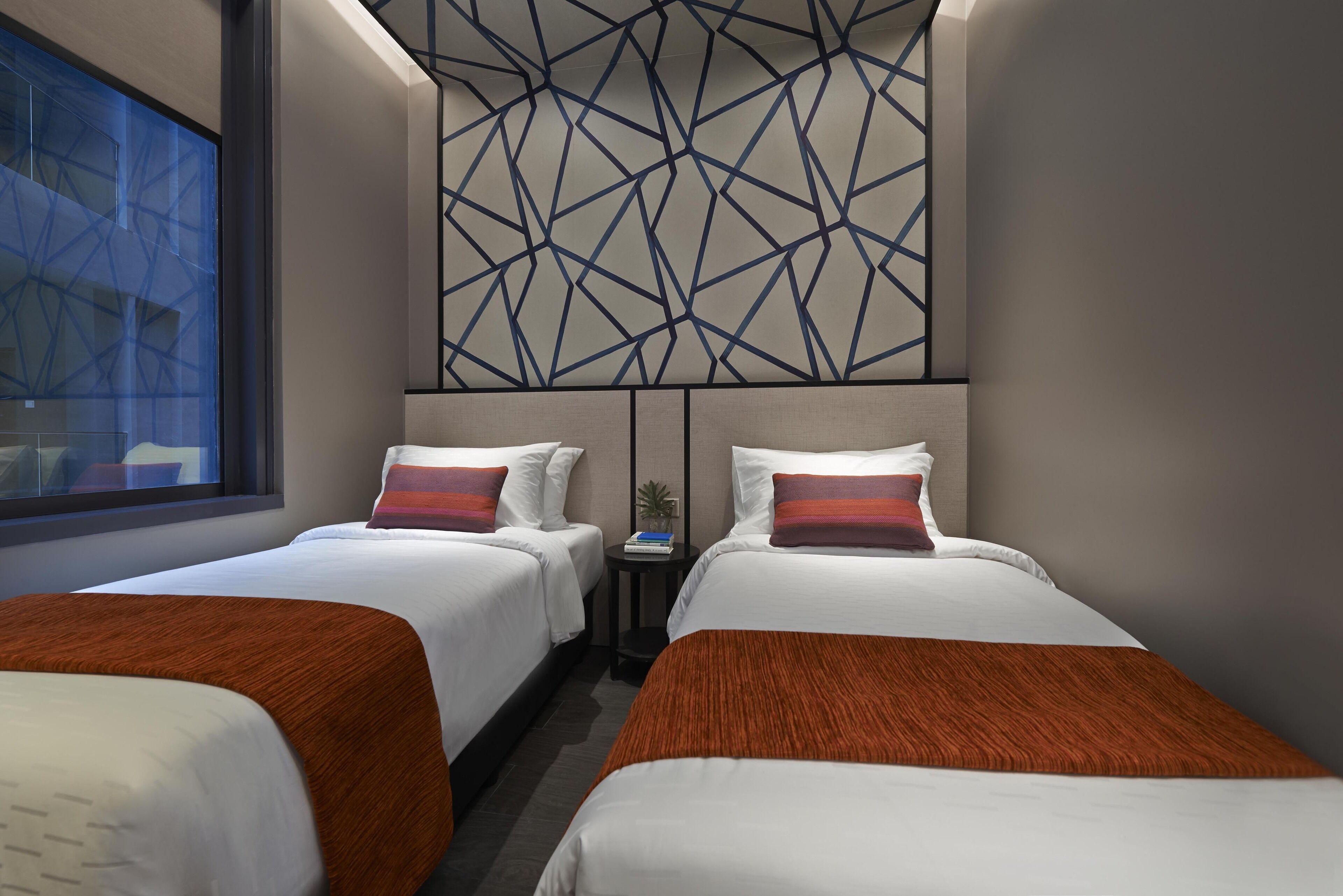 Habitación de hotel con dos camas, diseño geométrico en la pared y una mesita con una planta.