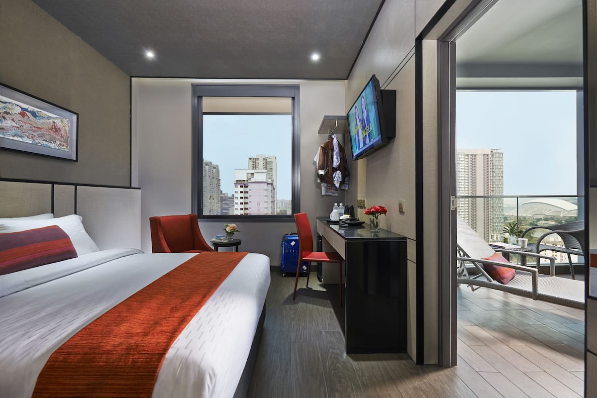 A modern hotel room with a large bed, a desk, a chair, a TV, and a balcony with a view of the city.