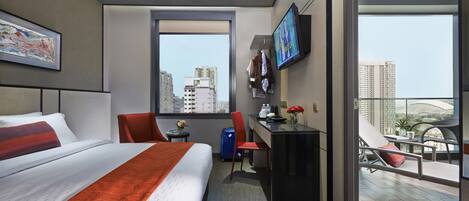 Habitación de hotel moderna con una cama grande, un escritorio, una silla, un televisor y un balcón con vistas a la ciudad.