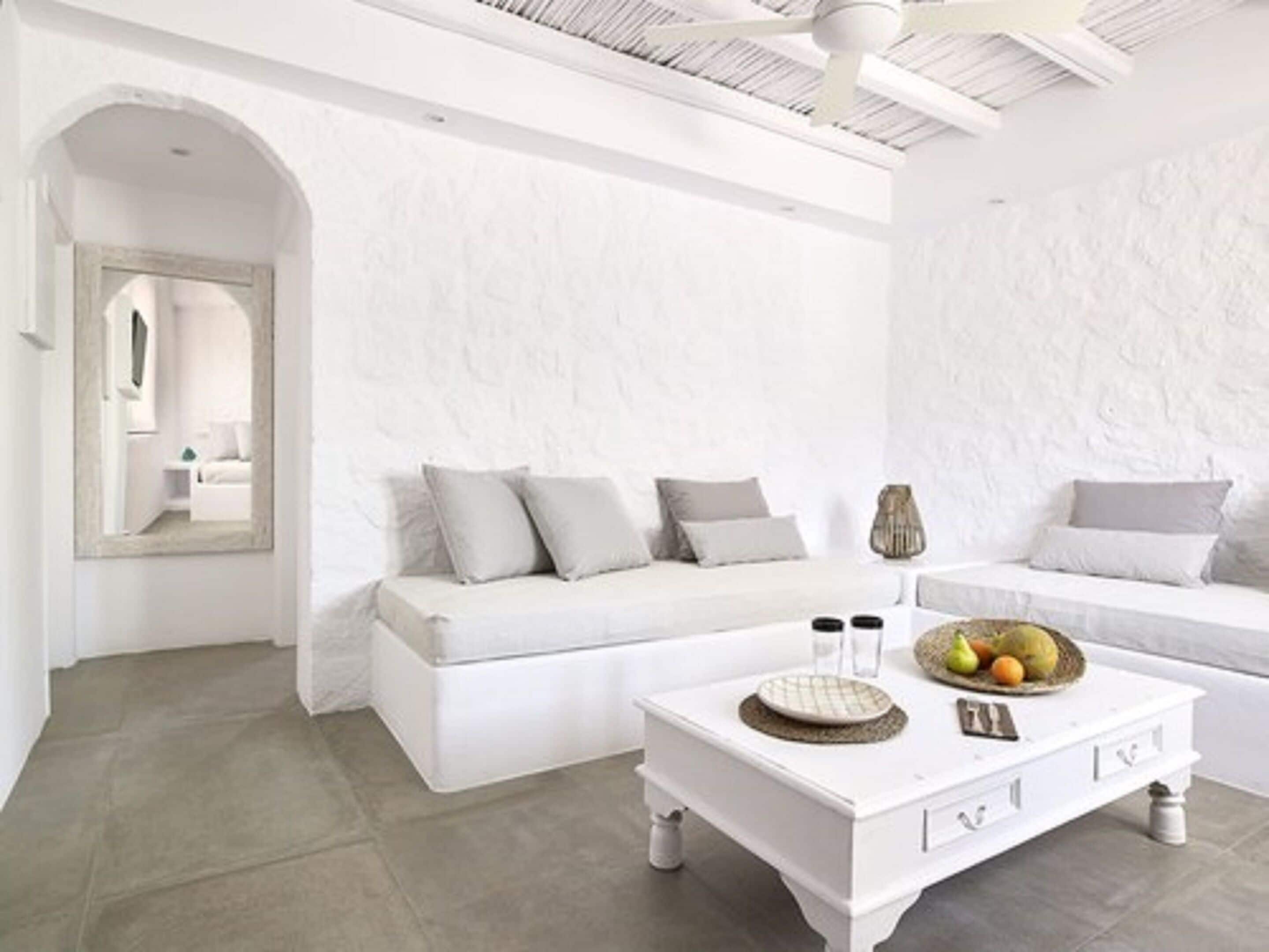 A modern, white-themed living room with a sofa, a coffee table with fruits, and a mirror reflecting the room.