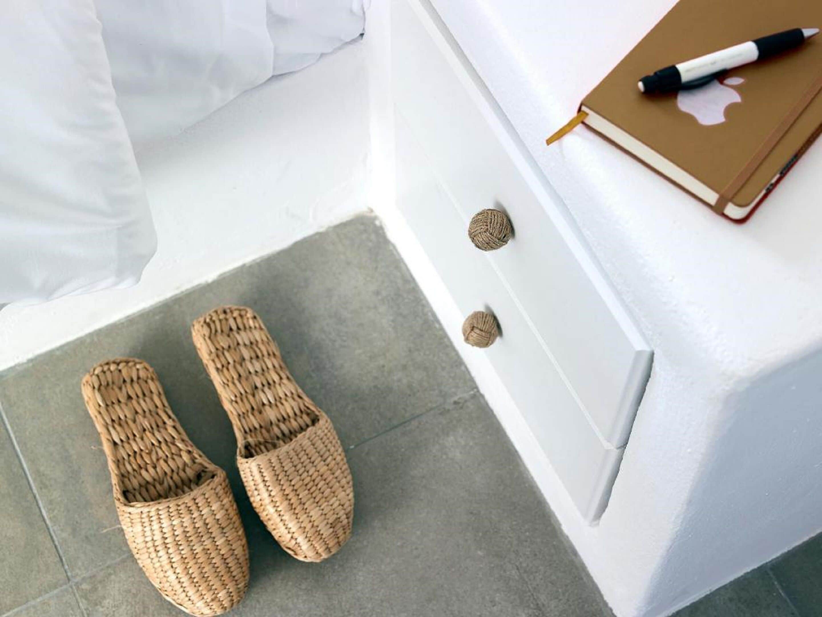 A room with a bed, a pair of woven slippers, a notebook, and a pen on a white dresser.