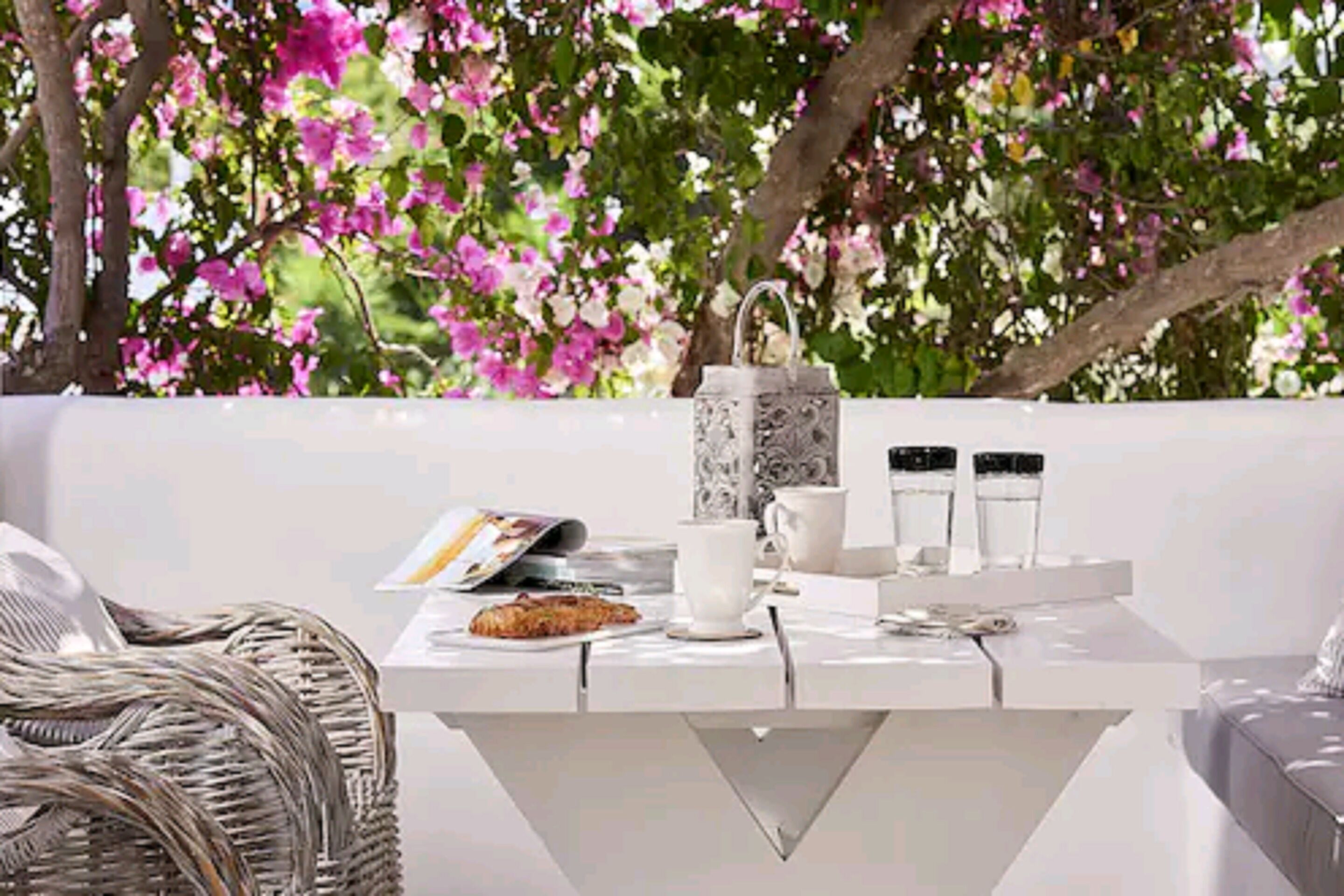 A white table with a magazine, a cup, and a plate with food, set against a backdrop of pink flowers and a tree.