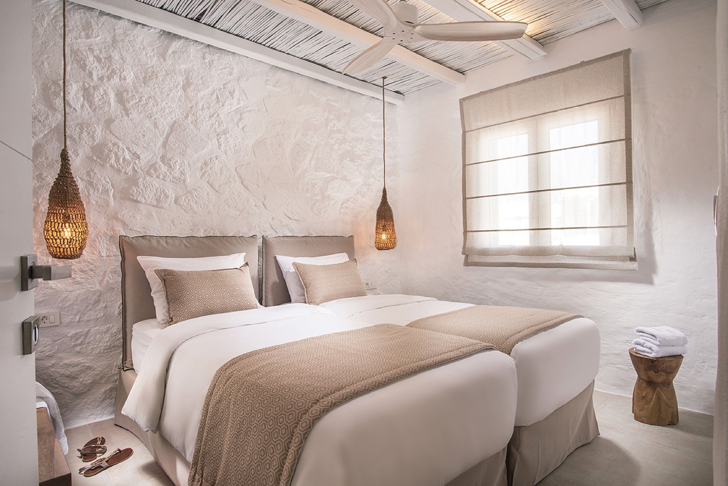 A bedroom with a large bed, two hanging light fixtures, a window with blinds, and a small stool with towels.
