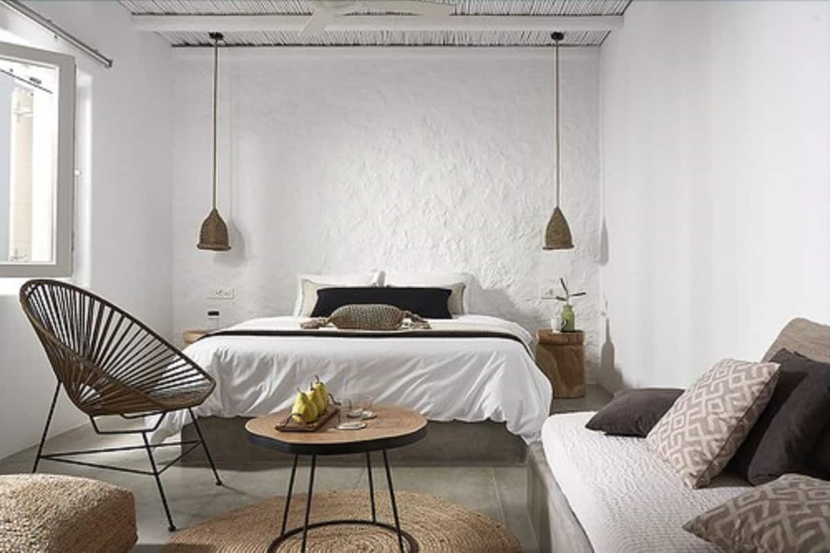 A minimalist bedroom with a bed, two pendant lights, a chair, a small round table, and a bedside plant.