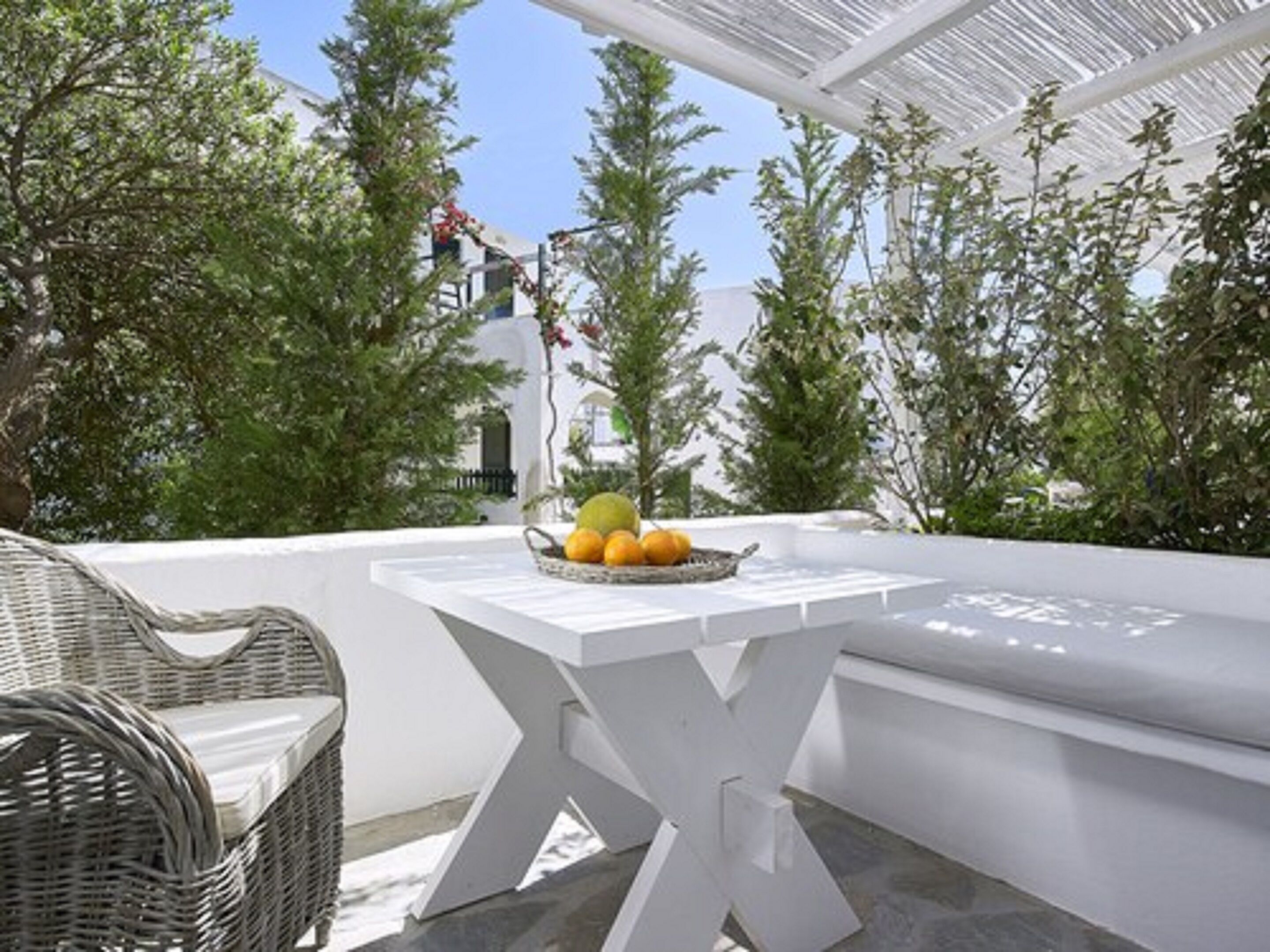 Un espace extérieur aménagé avec des sièges blancs, comprenant une chaise en osier et une table sur laquelle est posé un panier de fruits.