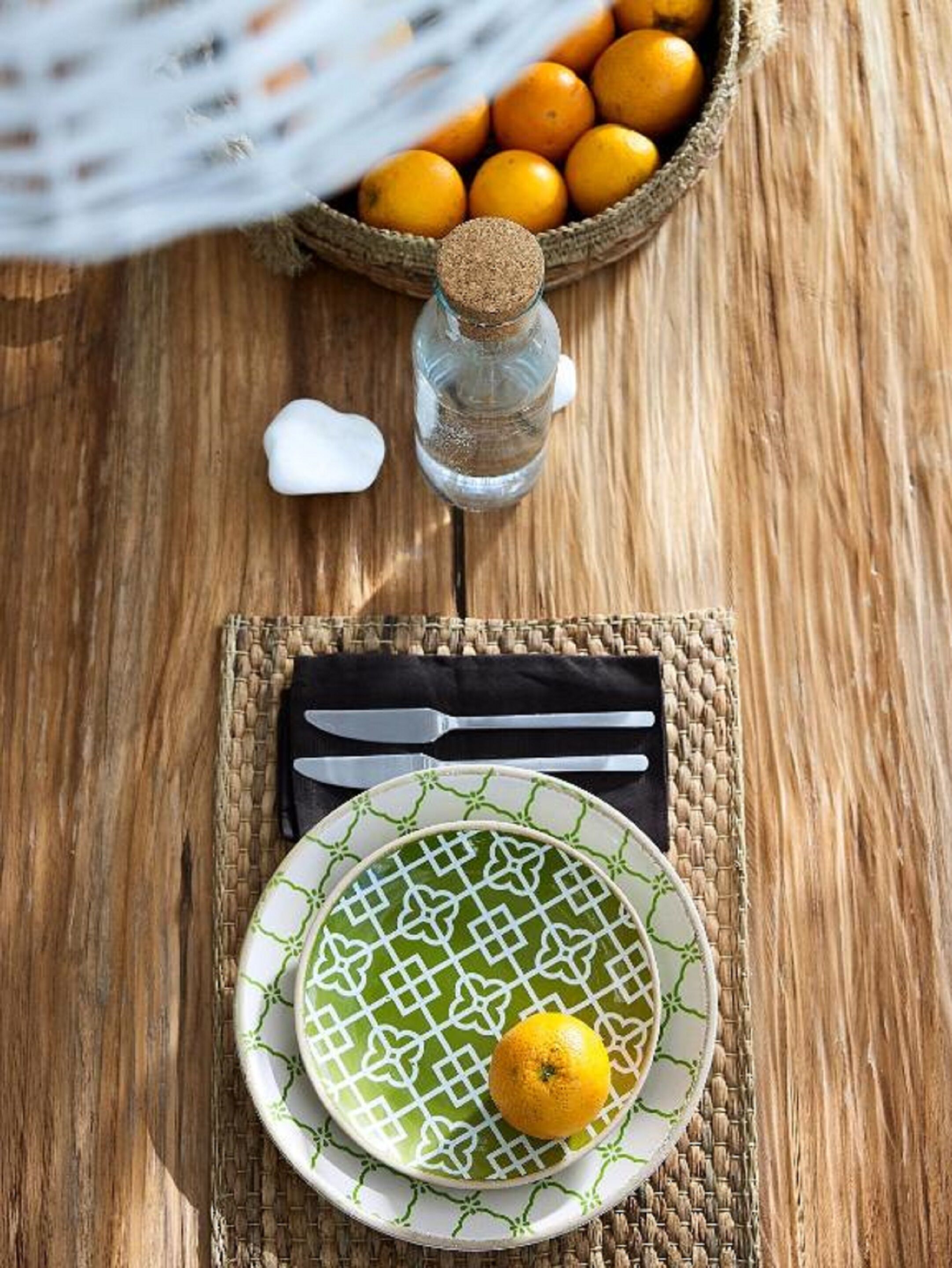 Una mesa de madera con un posavasos tejido, un plato con un diseño verde, un limón, una botella y una cesta de naranjas.