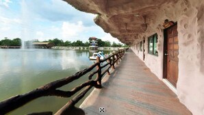 A wooden walkway beside a river with a rustic building and a fountain in the background. - Chart Thip Resort (Sung Noen)