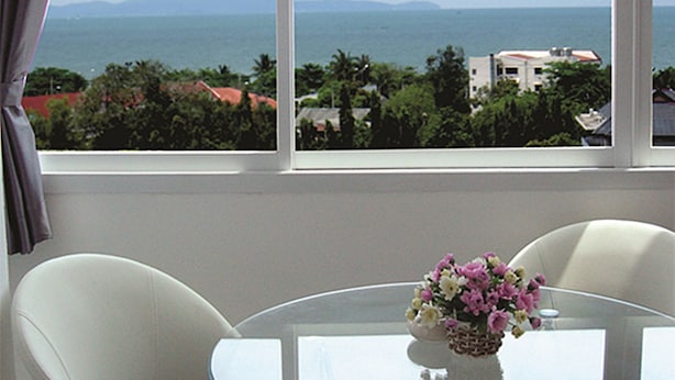 A room with a round glass table, two white chairs, a vase of flowers, and a view of the sea and mountains through large windows. - Grand Residence Jomtien (Pattaya)