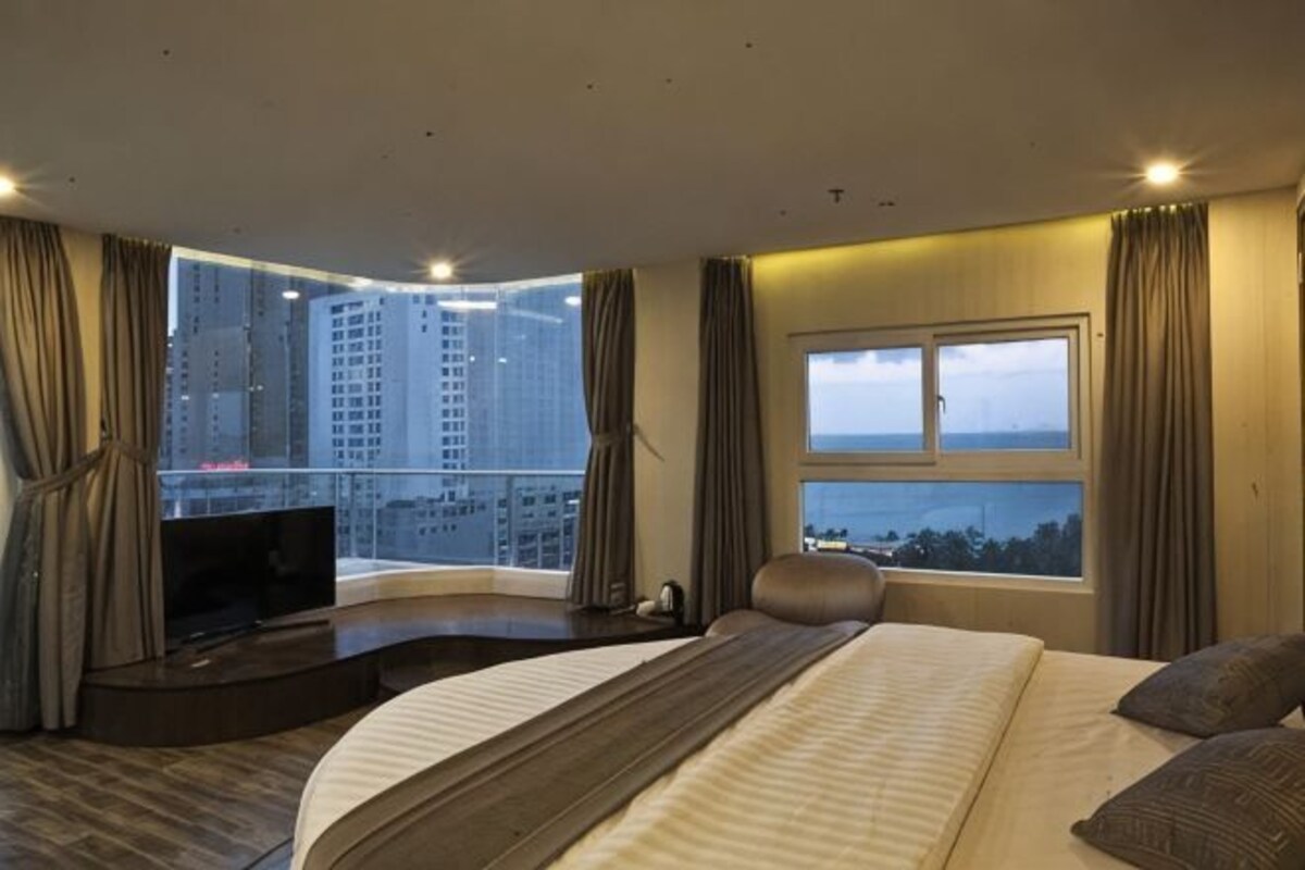 Habitación de hotel con una cama grande, televisión, balcón con vistas a la ciudad y un ventanal amplio.