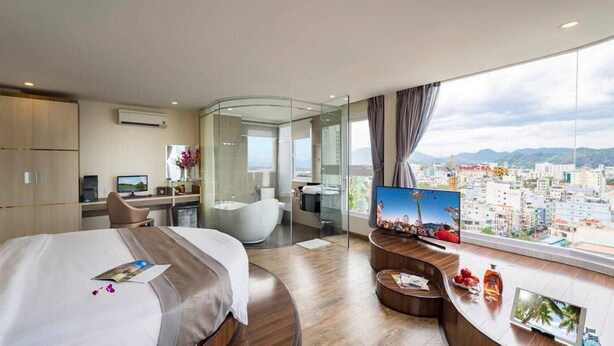 A modern hotel room with a large bed, a desk with a computer, a glass-walled shower area, and a TV displaying a cityscape. - New Sun Hotel (Nha Trang)