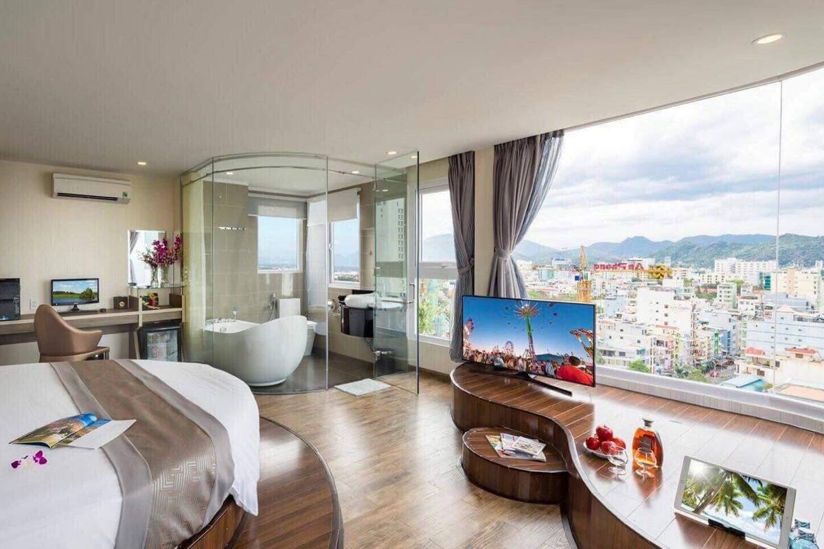 Une chambre d’hôtel moderne dotée d’un grand lit, d’un bureau avec un ordinateur, d’une douche à parois de verre et d’un téléviseur diffusant des images de la ville.