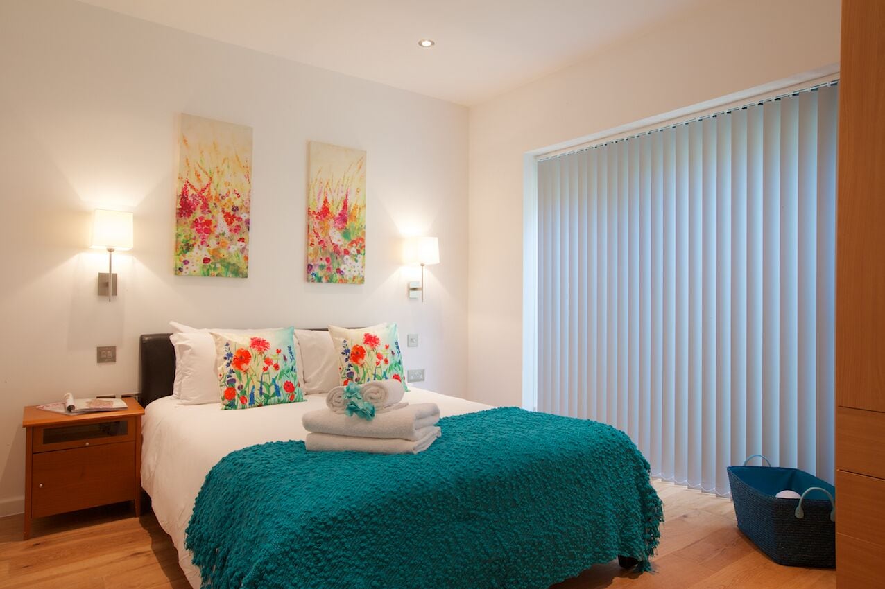 A bedroom with a bed, two wall-mounted lamps, a nightstand, a window with blinds, and a decorative wall piece.